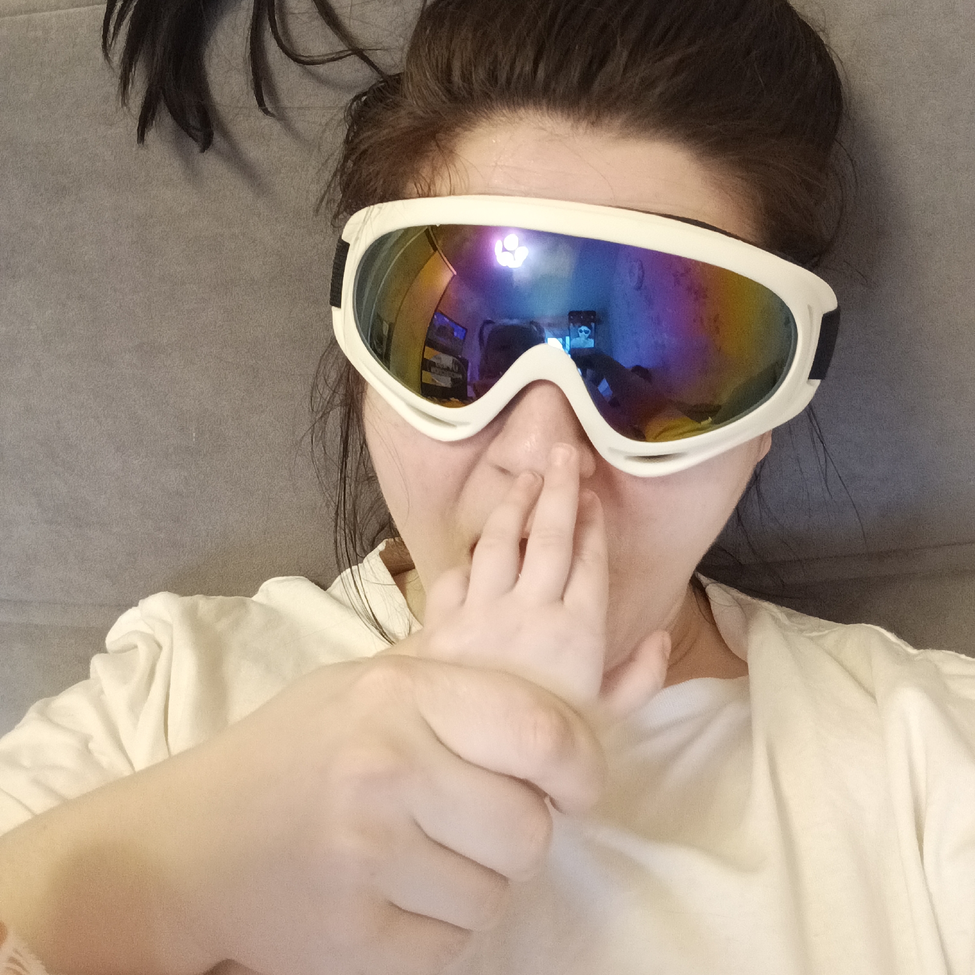 A woman is lying on a gray surface wearing large, white-framed goggles with rainbow-colored lenses. The woman has brown hair pulled back, with strands escaping. A small child's hand is near her face, with fingers touching the woman's nose. The woman is wearing a white shirt. The reflection in the goggles' lenses shows a room interior with electronic devices, and a person wearing the same goggles. The location is Chaikovsky, Russia.