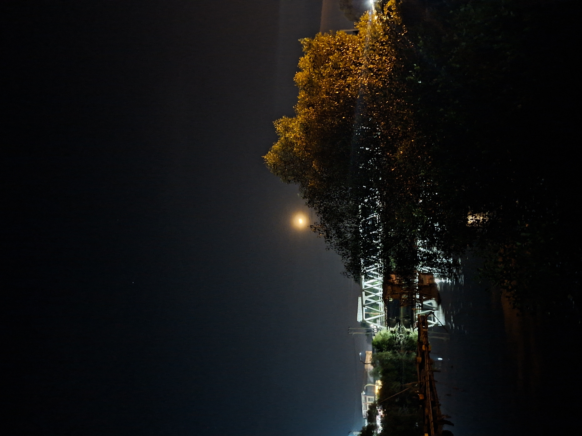 The image shows a night scene in Lhokseumawe, Indonesia. A brightly lit tower structure stands near a tree with dense foliage. A light source, possibly the moon, is visible in the dark sky. The scene is overall dark with points of illumination.