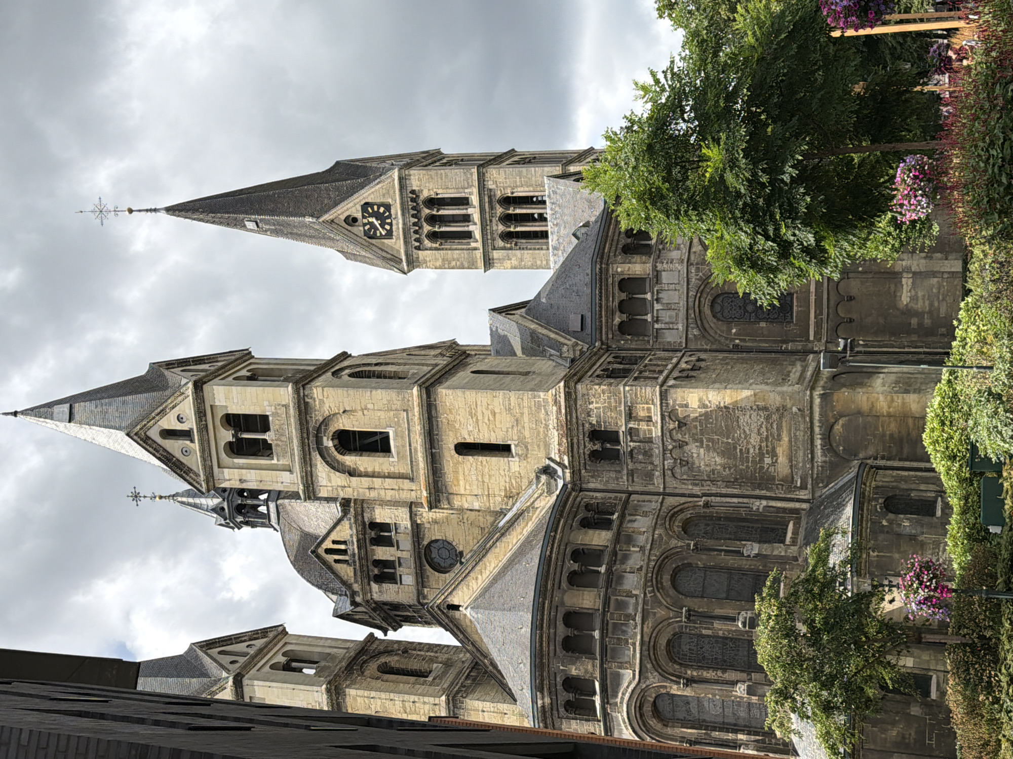 The image shows the exterior of the Saint Christopher Cathedral in Roermond, Netherlands. It is a large, imposing stone structure with multiple spires and towers. The architecture is Romanesque and Gothic in style, with arched windows and intricate stone carvings. The cathedral is surrounded by lush green trees and flowering bushes. The sky is overcast, suggesting a cloudy day. There are no people visible in the image. The perspective is from a low angle, looking up at the cathedral.