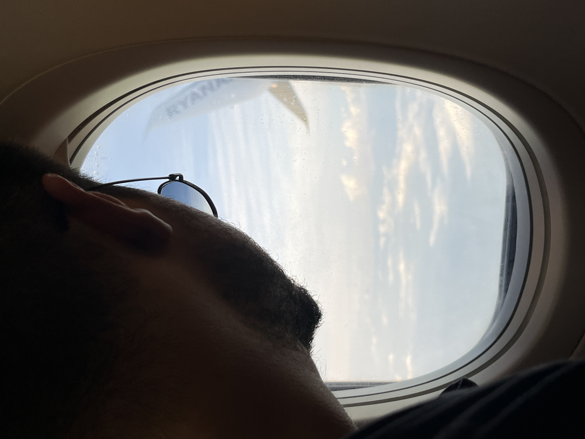 The image captures a close-up, eye-level view from inside an airplane cabin, depicting a man looking out through a circular window. The man, seen from the back of his head and right profile, has dark hair and a neatly trimmed short beard or stubble along his jawline. He is wearing a pair of dark-rimmed sunglasses, with one temple resting against his ear. His left shoulder, clad in dark clothing, is partially visible in the lower left. The airplane window frame is a prominent feature, showing light beige interior paneling. Through the window, a bright sky with scattered, fluffy white clouds is visible below, suggesting the plane is at a significant altitude. A portion of the aircraft's wing is seen in the upper right section of the window, with the faint branding "RYANAIR" visible on its surface. The window pane itself shows minor condensation or dirt. The scene takes place aboard a Ryanair aircraft, with the flight potentially originating from, destined for, or flying over Sofia, Bulgaria.