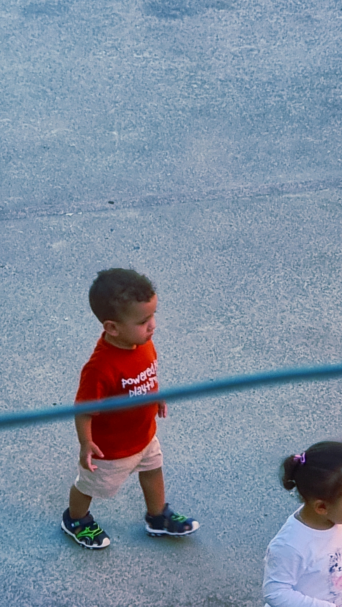 A young boy with dark curly hair and wearing a red t-shirt with white lettering and tan shorts walks across a grey, textured surface. The lettering on his shirt appears to read "Powered by Play." He is wearing dark sandals with green accents. To the right and slightly behind him, the back of a young girl's head is visible, with dark hair pulled back with a purple hair tie. She is wearing a white long-sleeved shirt. They are on a paved surface that appears to be outdoors, possibly a playground or park area in Sant Joan Les Fonts, Spain. A thin, blurred blue line runs horizontally across the lower portion of the image.