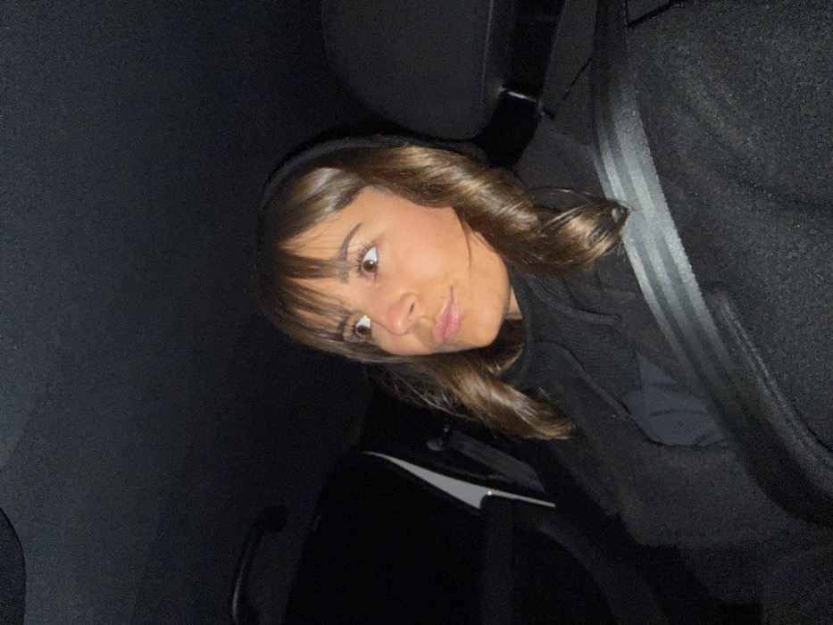 A young woman with bangs and brown hair sits in a car, wearing a seatbelt. She looks towards the camera with a slight pout. The interior of the car is dark, with only the woman's face and part of the seat visible. The lighting suggests it's nighttime.