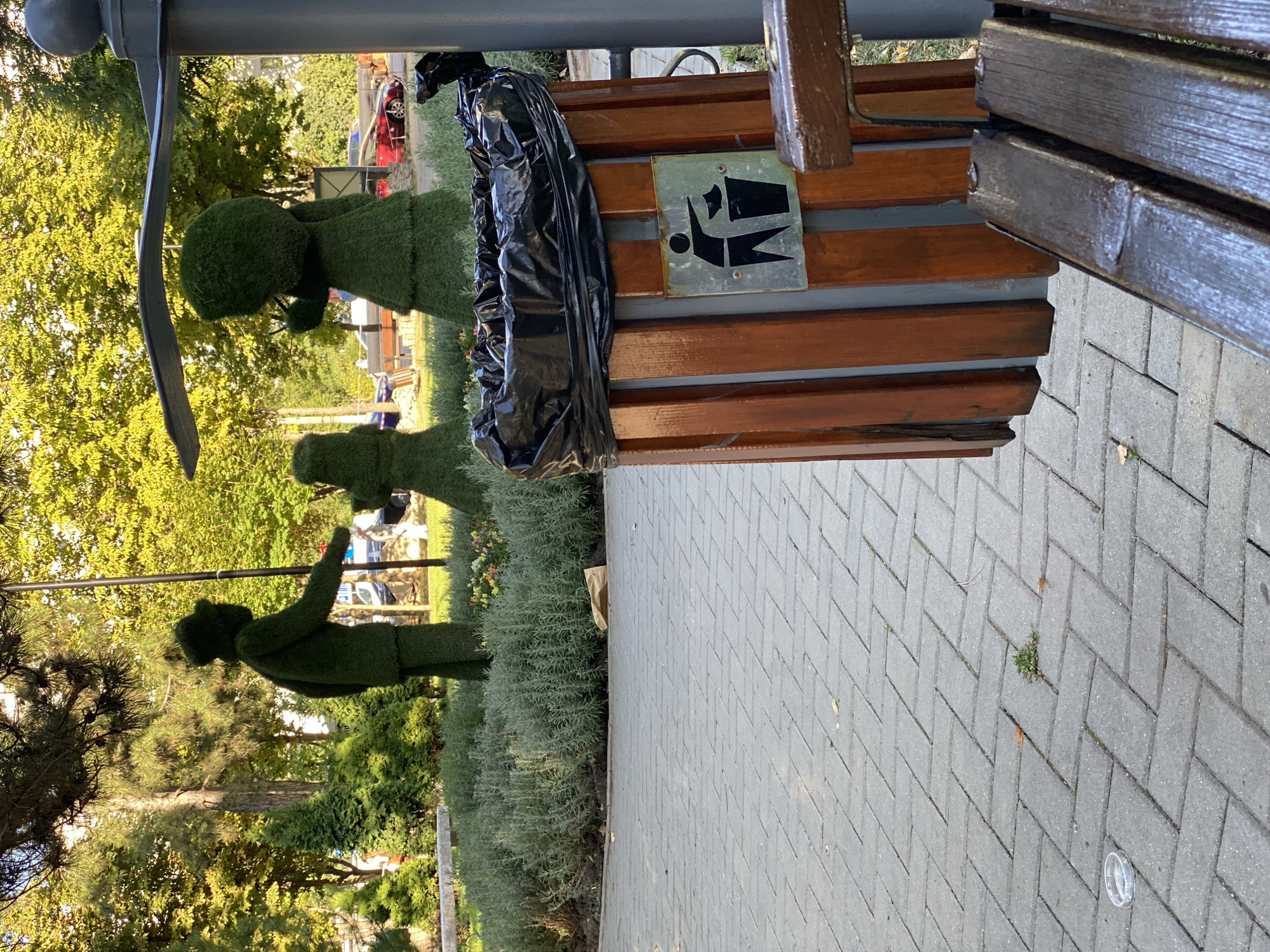 The image shows a park scene in Chișinău, Moldova. In the foreground, a paved walkway made of gray rectangular bricks is visible. To the right, a wooden trash bin with a black plastic liner is positioned next to a park bench. The trash bin has a metal sign with a black silhouette indicating proper waste disposal. In the background, there are topiary figures shaped like people, one appearing to be an adult with a child. These figures are made of dense green foliage. Beyond the topiaries, a row of low-lying green bushes and some flowering plants are present. Trees with lush green foliage form the backdrop of the scene, with sunlight filtering through the leaves. A light-colored pole with a curved canopy extends from the right side of the frame, partially obscuring some of the topiary figures. The overall environment suggests a well-maintained public park.