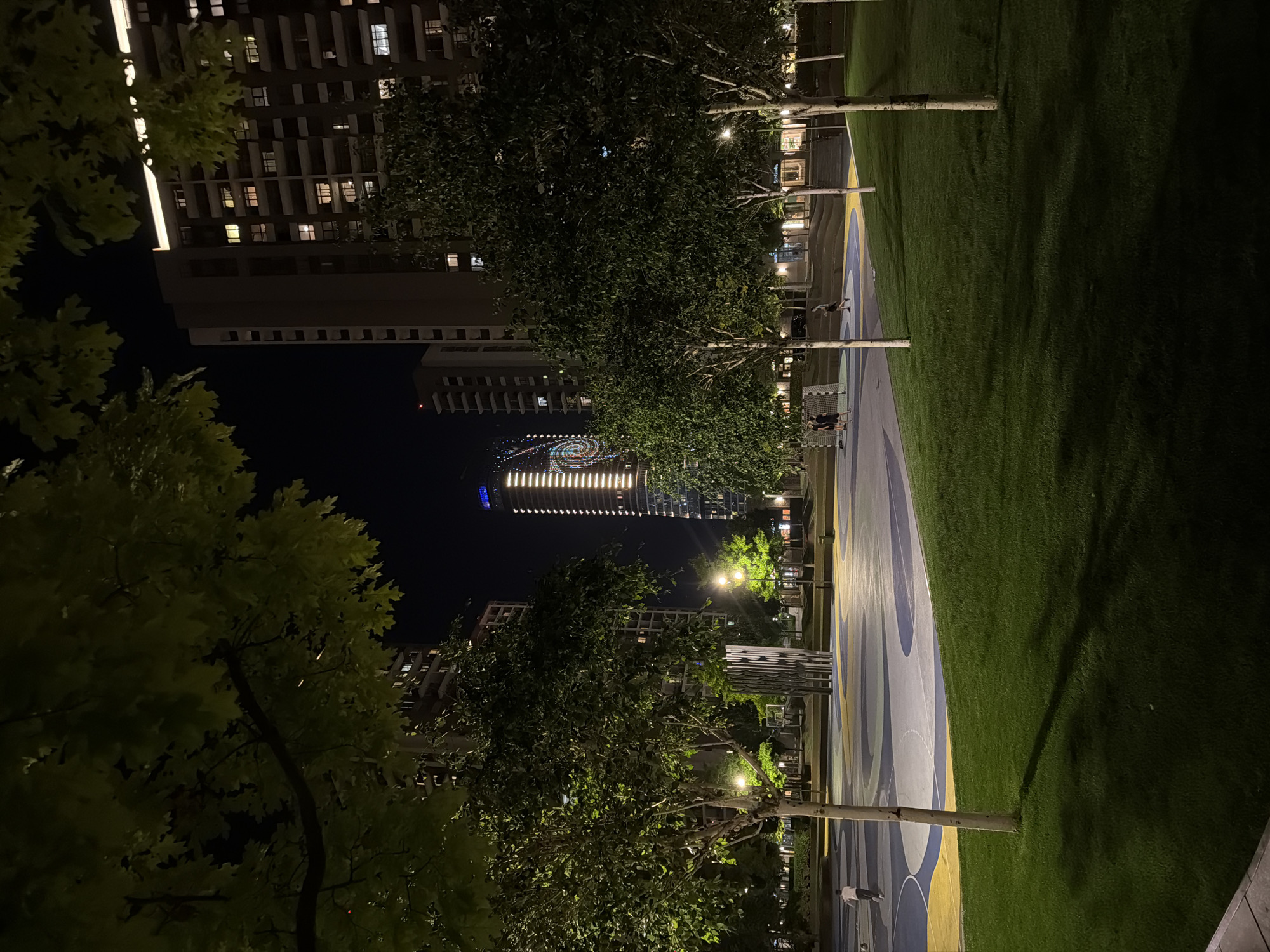 The image shows a nighttime scene in Belgrade, Serbia. A path, painted in shades of blue and yellow, runs alongside a patch of green grass. Trees with illuminated foliage frame the scene on both sides. In the background, illuminated buildings rise into the night sky. The lights of a high-rise building create a distinctive, circular pattern. People are walking on the path. A building complex with visible windows is on the left.
