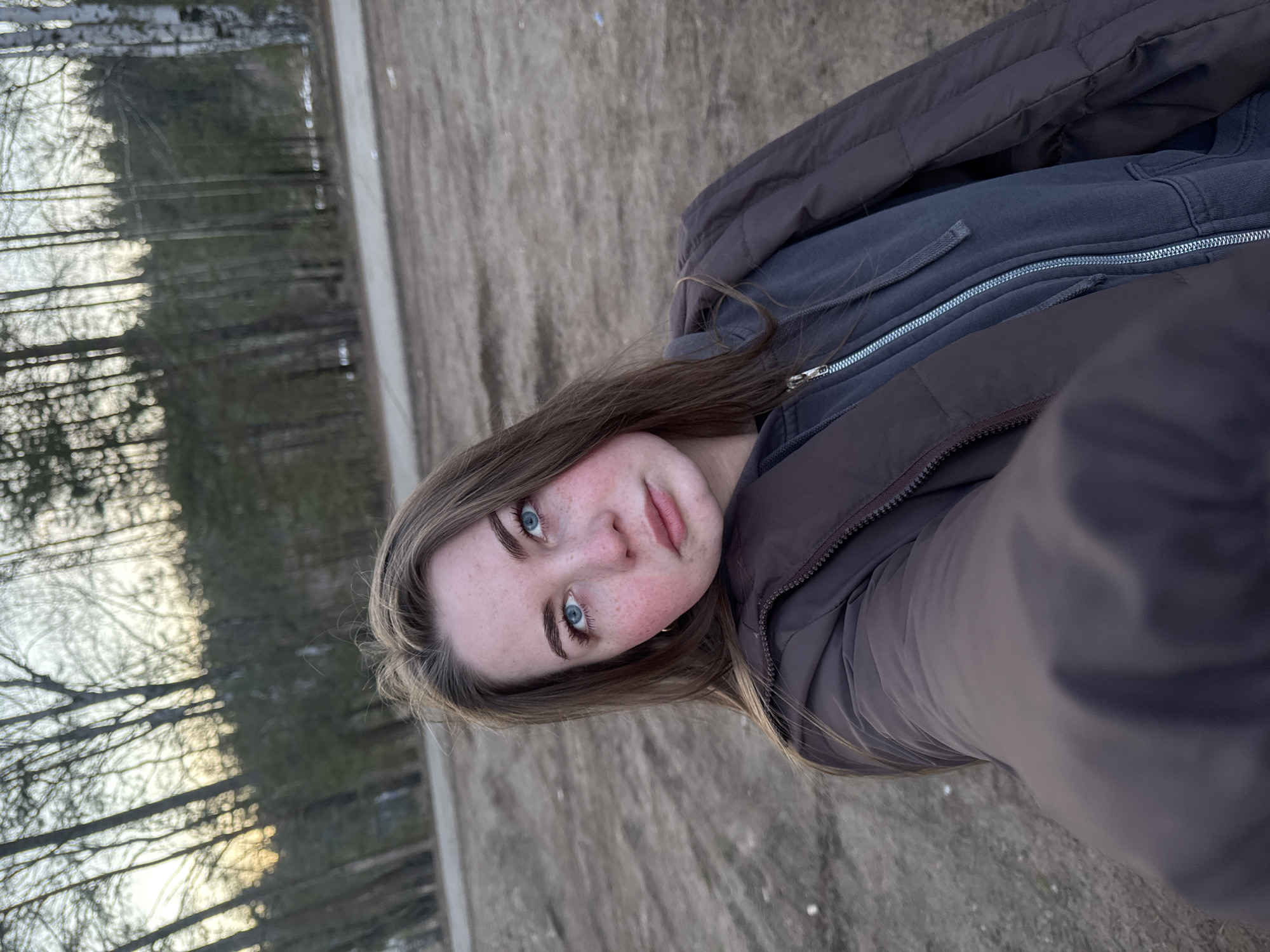 A person is centered in the frame in an outdoor setting. The person has fair skin, brown hair, and blue eyes with noticeable freckles on their cheeks and nose. They are wearing a dark gray zip-up jacket under a brown puffy coat. The background appears to be an outdoor area with trees visible to the left and a ground surface that looks like dirt and possibly some sparse grass. The photo is taken outdoors, likely in Noviy, Russia. The overall image has a cool, natural light.