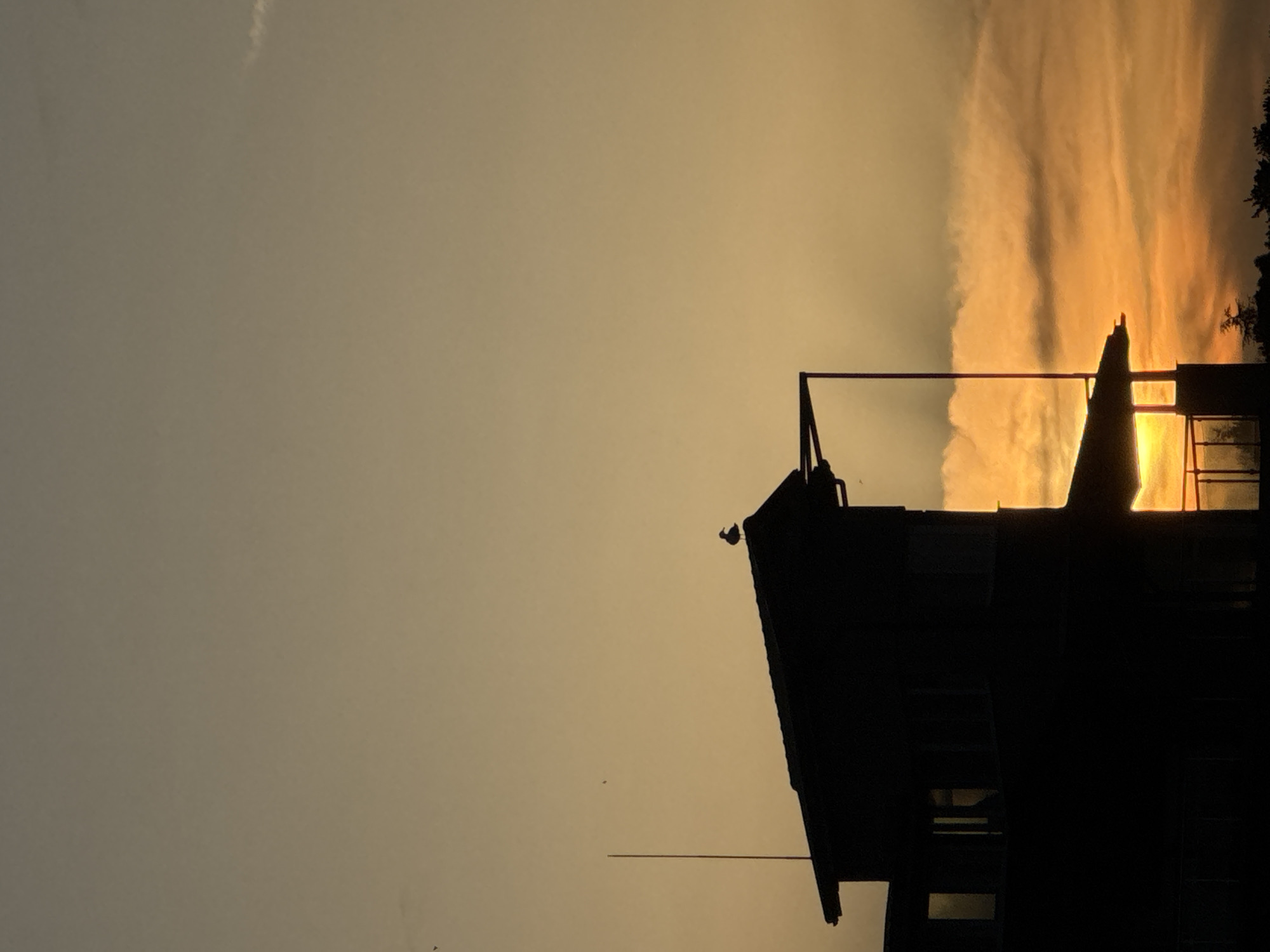 The image shows a silhouette of a building against a sunset sky. The building is dark and detailed, with multiple levels and a peaked roof. A flag pole is visible extending from the side of the building. The sky is a gradient of colors, with the brightest orange and yellow hues concentrated near the horizon. A light, wispy cloud is near the top left. The image is taken in Tsarevo, Bulgaria.