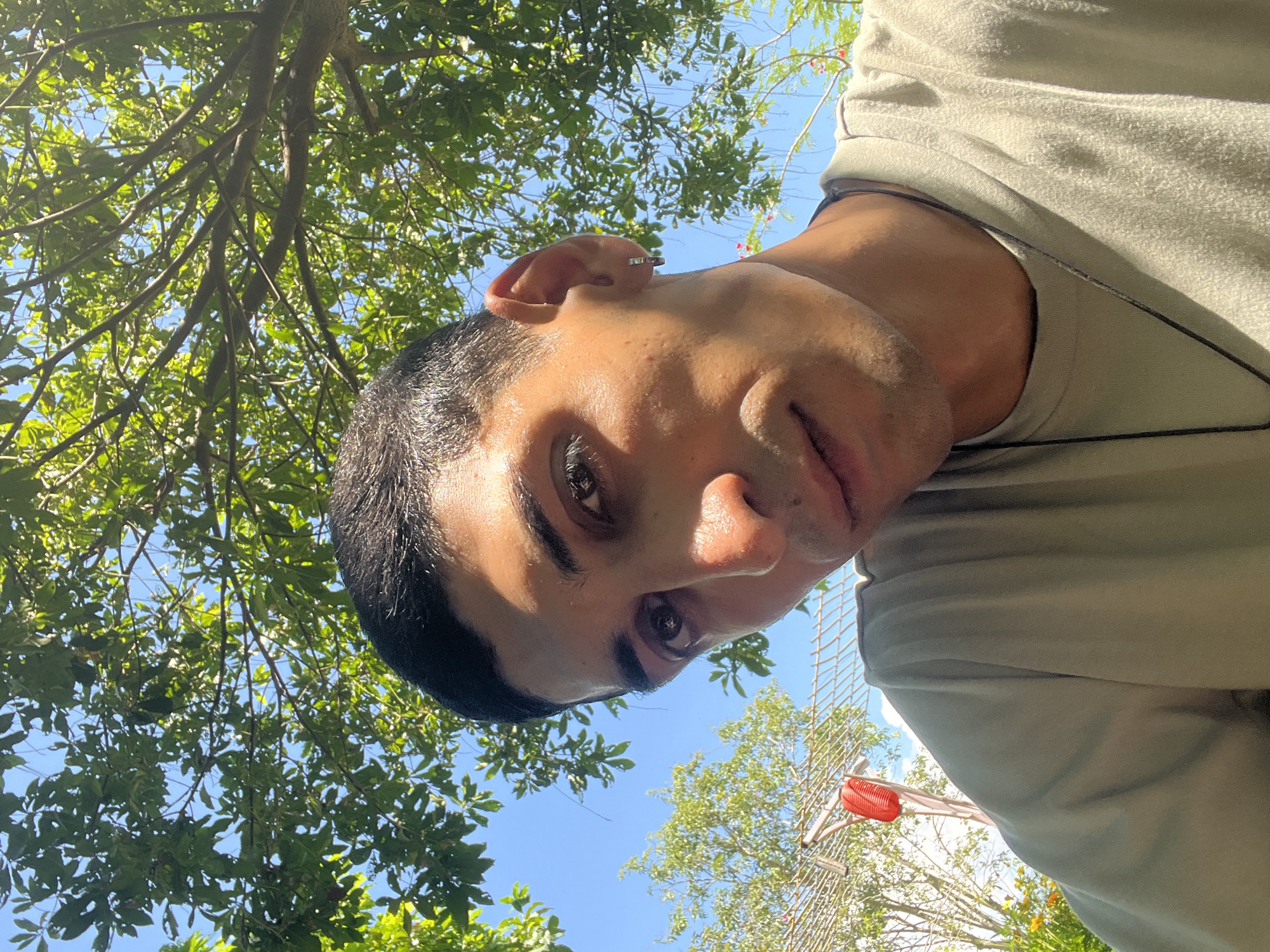 A young man with dark hair and brown eyes is pictured in a close-up, low-angle shot, looking directly at the camera. He is wearing a light-colored t-shirt and a dark necklace. He has a small silver hoop earring in his left ear. The background is filled with lush green tree leaves and a bright blue sky. In the upper left corner of the image, there is a glimpse of a red lantern hanging from a structure, and some yellow flowers are visible near the bottom left. The lighting suggests it is daytime, possibly outdoors in a garden or park-like setting. The context of the image, including the specific location in Nam Cam Ranh, Vietnam, is not discernible from the visual information alone.