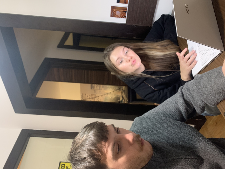 A man and a woman pose for a photo. The man, facing the camera, wears a gray sweater. Behind him, a woman with long blonde hair, clad in a black hoodie, sits at a laptop, her hand hovering over the keyboard. A framed image of a bridge hangs on the wall, partially visible through a doorway. The scene is captured at an angle, slightly tilted, with a second doorway visible.