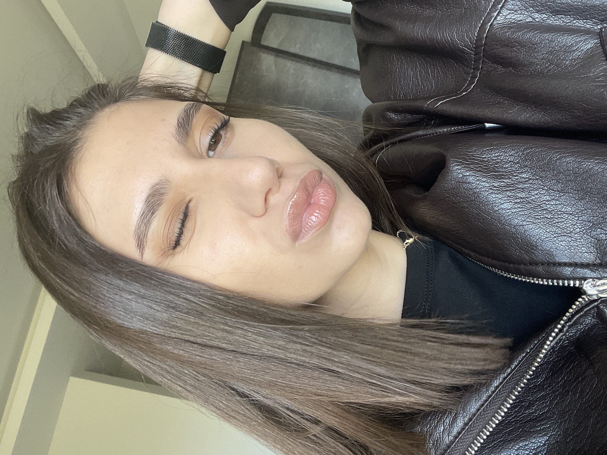 A young woman with long, brown hair, light skin, and brown eyes is posing for a selfie. She is wearing a dark brown leather jacket and a black shirt underneath. A black watch band is visible on her wrist. The background is slightly out of focus, showing a wall and a piece of furniture. There is no discernible information to indicate that this image was taken in Sofia, Bulgaria.