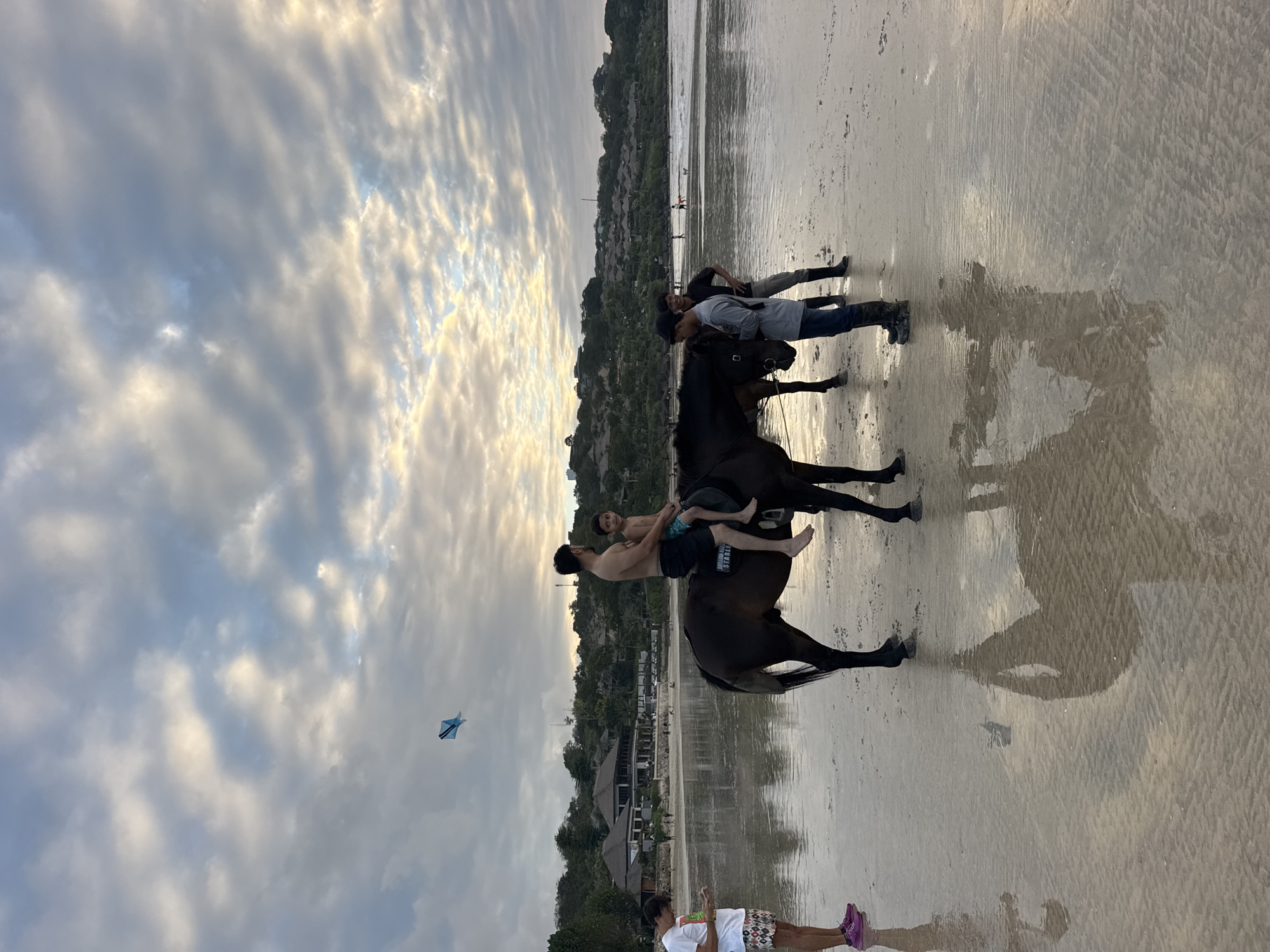 A horse is being led by two men on a sandy beach in Kuta Selatan, Indonesia. A man and a young boy are seated on the horse, with the man appearing to be talking to the boy. A woman in a white t-shirt and patterned shorts stands to the left, looking at her phone. The sky is cloudy and the sun is setting, casting reflections on the wet sand. In the background, there are buildings and trees, and the ocean is visible. A blue kite is flying in the sky.