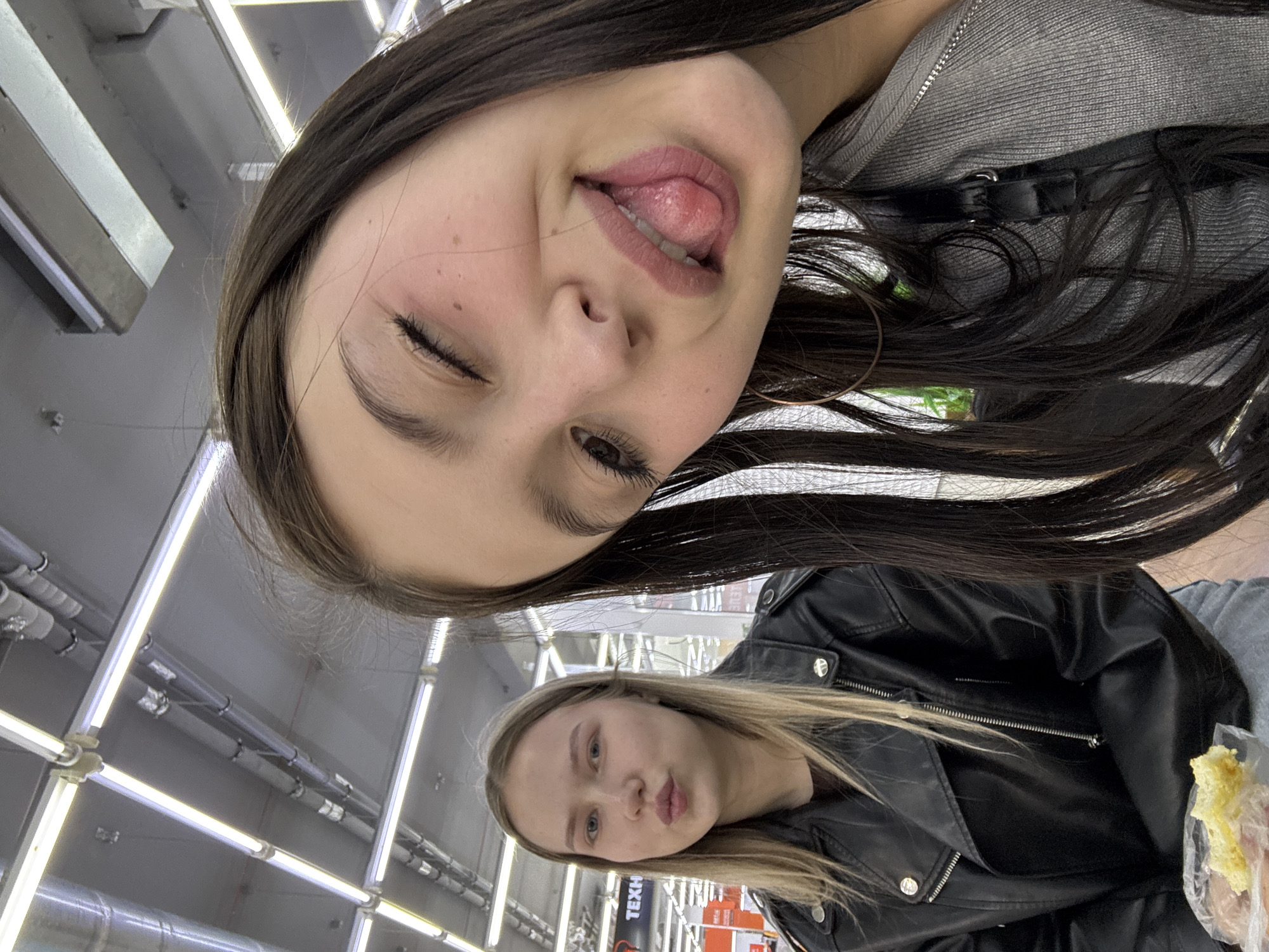Two young women are photographed indoors. The woman in the foreground, on the right, is closer to the camera and is looking down, sticking her tongue out and winking. She has long, dark hair and is wearing a grey zip-up jacket and large hoop earrings. The woman in the background, on the left, is looking forward with her lips pursed. She has long, blonde hair and is wearing a black leather jacket. They appear to be in a public place, possibly a shopping center, indicated by the fluorescent lighting and shelving units visible in the background. The location is identified as Chaikovsky, Russia.