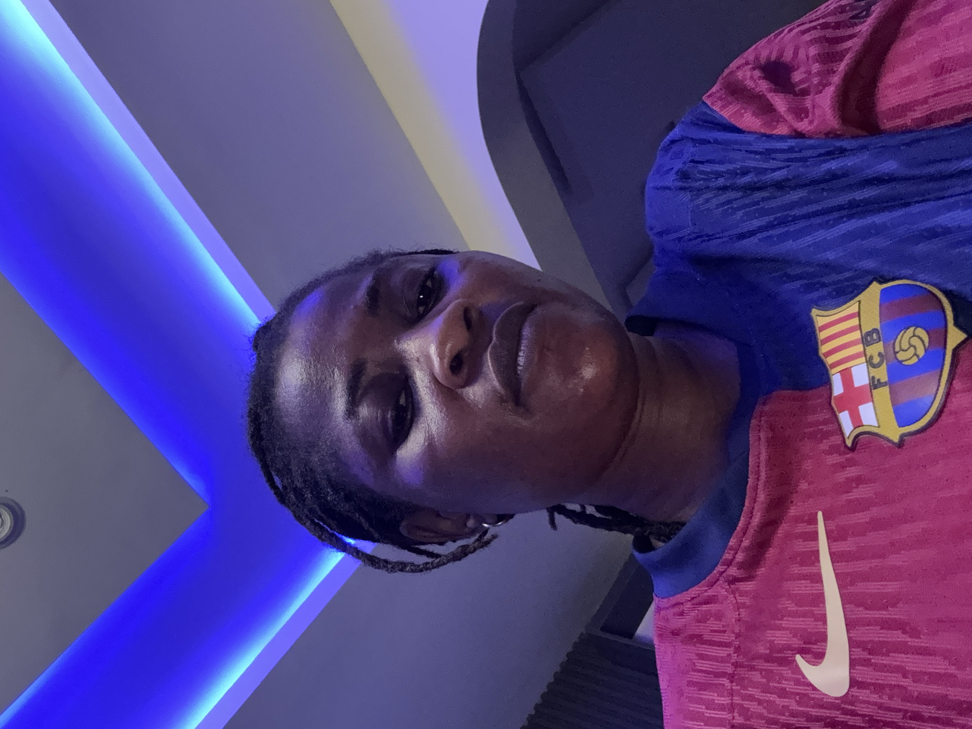 An overhead, medium shot captures a woman with dark skin and braided hair looking directly at the camera. She is wearing a FC Barcelona jersey, split between pink and blue. A Nike logo is visible on the pink side of the shirt. The background consists of a ceiling with blue LED lighting.