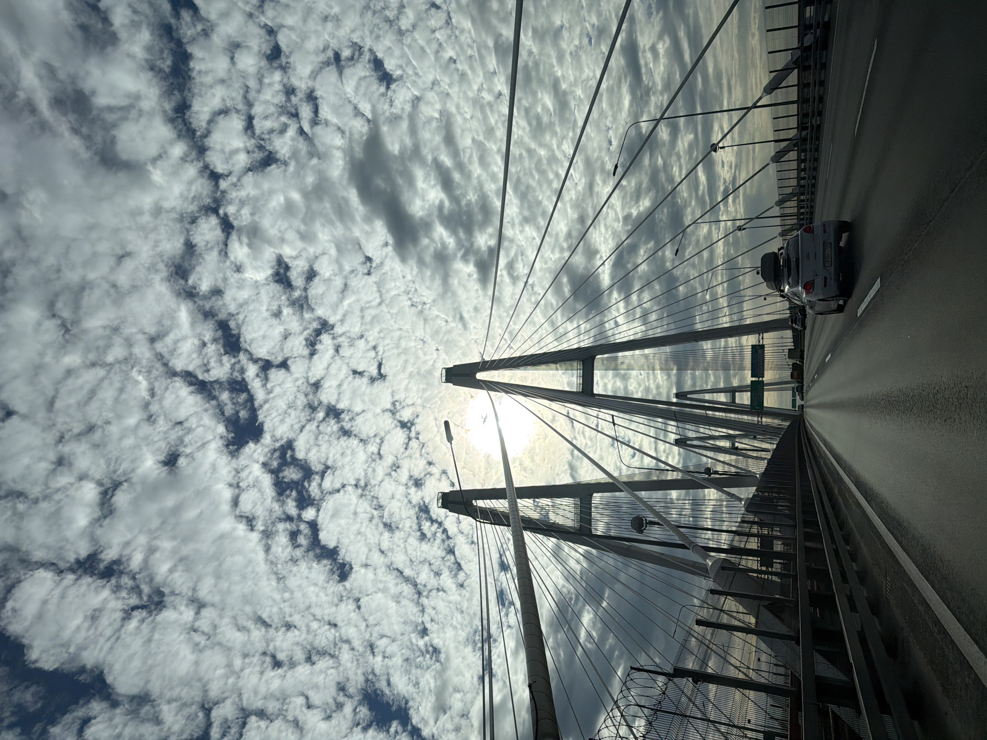 The image captures a view from a moving vehicle on a cable-stayed bridge, likely in an urban or coastal setting. The perspective is from the driver's seat, looking forward along the roadway. The bridge is characterized by its tall, twin towers and numerous cables supporting the deck. The sky above is filled with a dramatic display of cumulus clouds, with the sun peeking through, creating a bright, almost hazy effect.
In the foreground, the asphalt roadway is visible, showing lane markings and a slight sheen, suggesting it might be damp. A white car with a luggage rack on its roof is traveling in the right lane ahead of the viewer's vehicle. On the left side of the road, a metal fence with barbed wire on top runs along the edge of the bridge. Streetlights are mounted along the bridge structure. In the distance, other vehicles and traffic signs are discernible. The surrounding environment is not clearly visible, but the presence of water and what appear to be other structures can be inferred in the far distance, particularly on the right side of the bridge where the sky meets a darker horizon. The overall lighting suggests it is daytime, possibly late morning or afternoon.