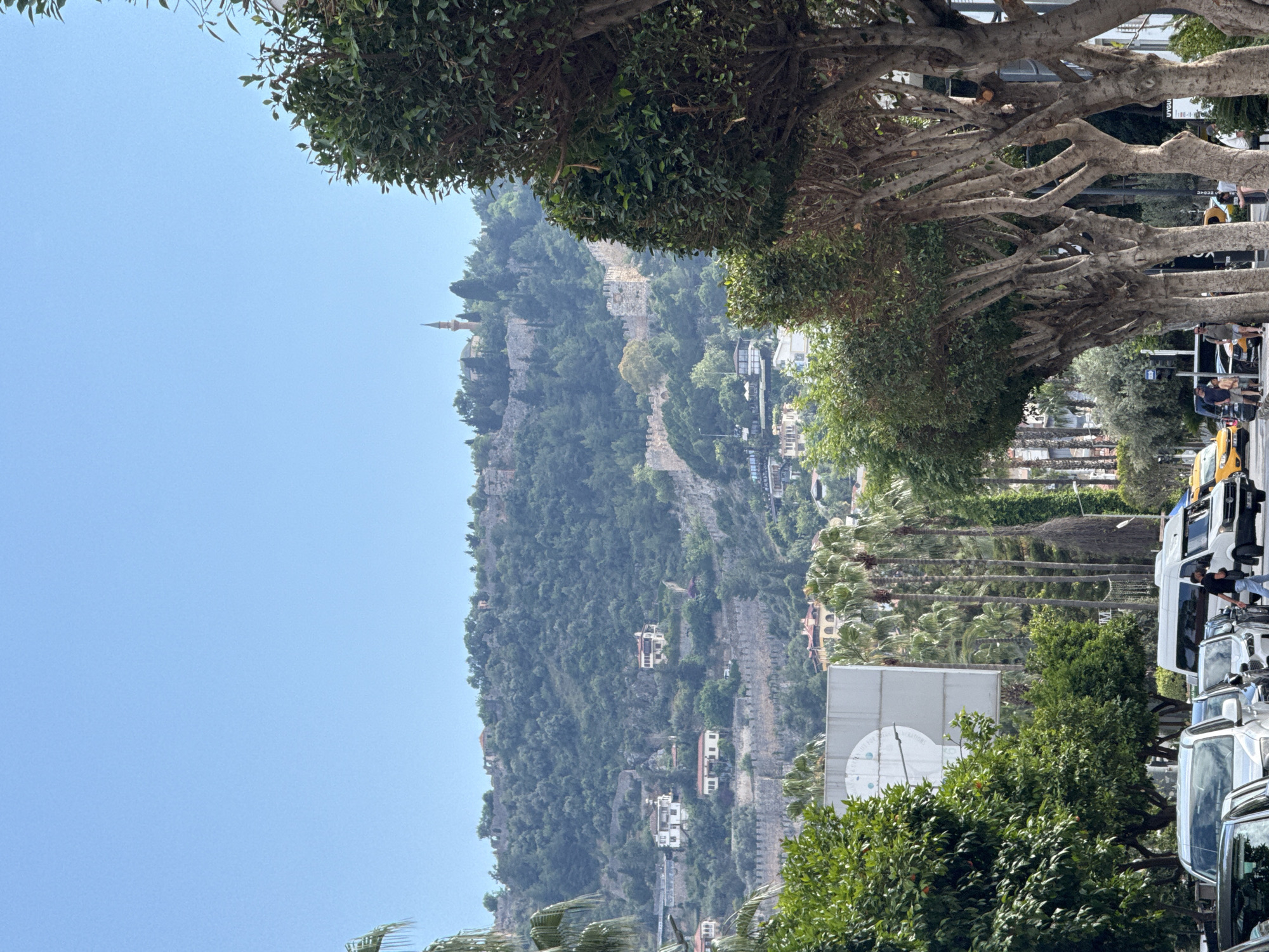 The image depicts a cityscape with a prominent hill or mountain in the background. A fortress or historical structure with a minaret is visible on the hilltop. The slopes are covered in dense vegetation, interspersed with buildings and architectural structures. The foreground reveals a street lined with trees, palm trees, and parked vehicles, including vans and taxis. People are seen walking along the street. The sky is clear and blue. The location is Alanya, Turkey, and the hill is topped by Alanya Castle.