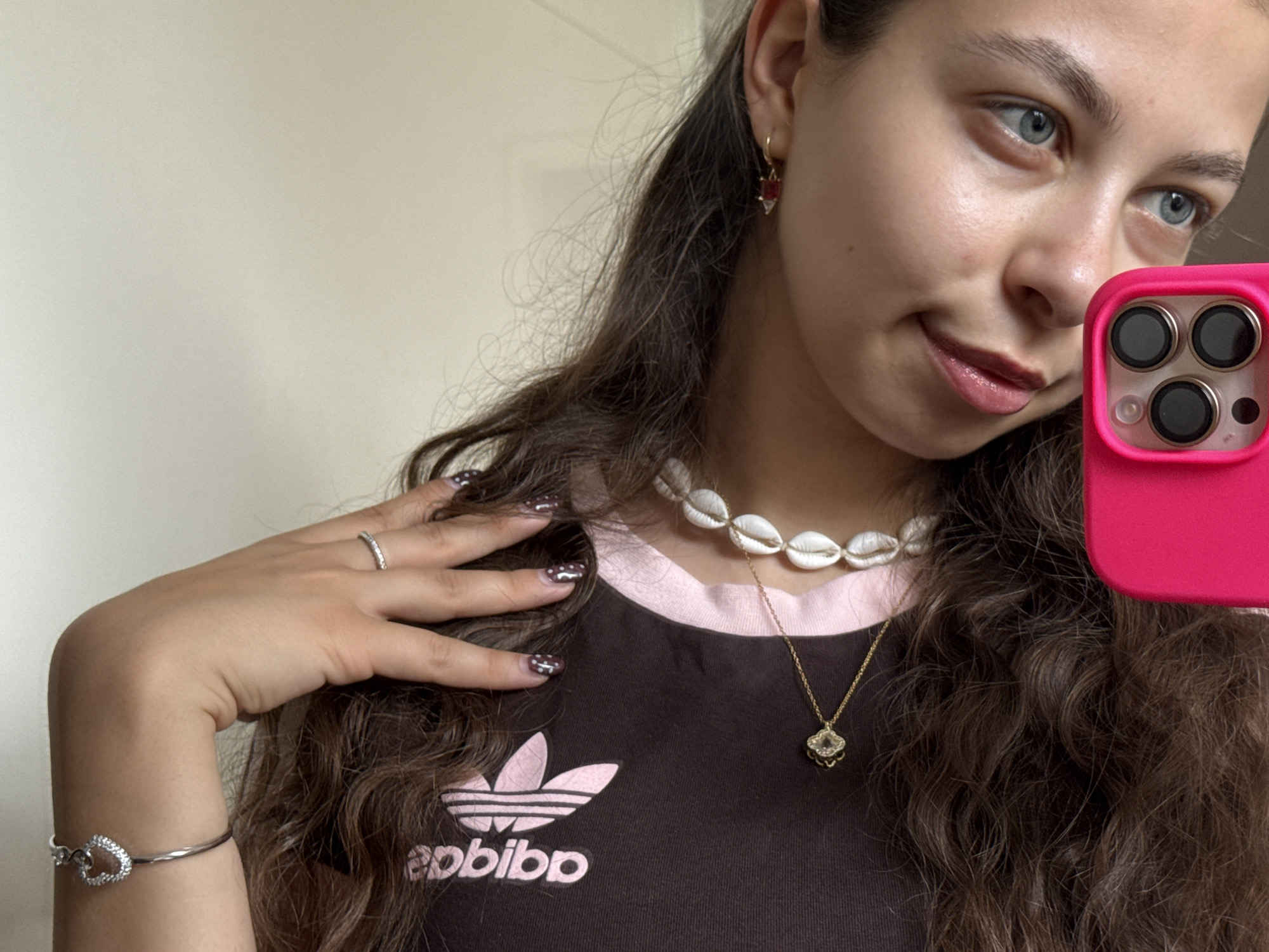 A young woman with long curly brown hair is taking a selfie. She is wearing a brown t-shirt with a pink Adidas logo, a seashell choker necklace, and a gold pendant necklace. She is also wearing earrings, a ring, and a heart-shaped bracelet. Her nails are painted with a white polka-dot design. The phone she is using to take the selfie is pink. The background is a plain, light-colored wall. There is no indication that this image was taken in Bruchköbel, Germany.