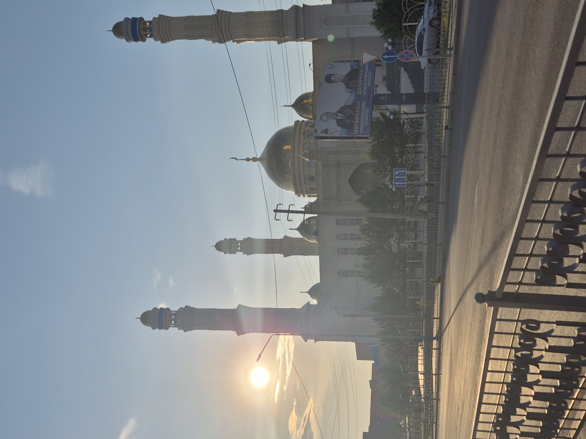 The image depicts a street scene in Kokshetau, Kazakhstan, featuring the Kenesary Mosque. The mosque has a main building with multiple golden domes and four minarets. A banner advertising "Shohibrmakov" is visible near the mosque. The street has a paved road with a metal fence on the side. The sun is setting, creating a bright glare in the lower left corner of the image. Power lines are strung above the mosque. A small vehicle is parked on the street, and traffic signs are visible.