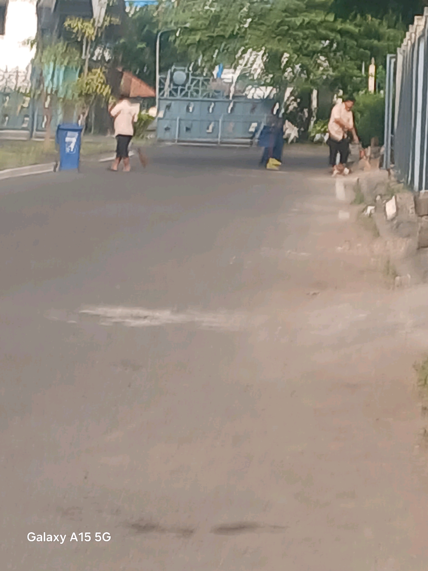 The image, taken in Surabaya, Indonesia, captures an outdoor scene with two individuals engaged in cleaning. On the left, a person in a light-colored shirt and dark pants is sweeping the paved ground near a blue trash bin with the number "7" visible on it. This individual is also holding what appears to be a brown umbrella over their head, suggesting it might be sunny or they are protecting themselves from debris. To the right, another person, also in a light-colored shirt and dark pants, is sweeping the ground and interacting with what seems to be a small pile of refuse. They are positioned near a grey fence or wall. The environment includes a road or pathway, trees in the background, and some industrial-looking structures or gates. The overall impression is of daily maintenance work being performed in an urban or industrial area.