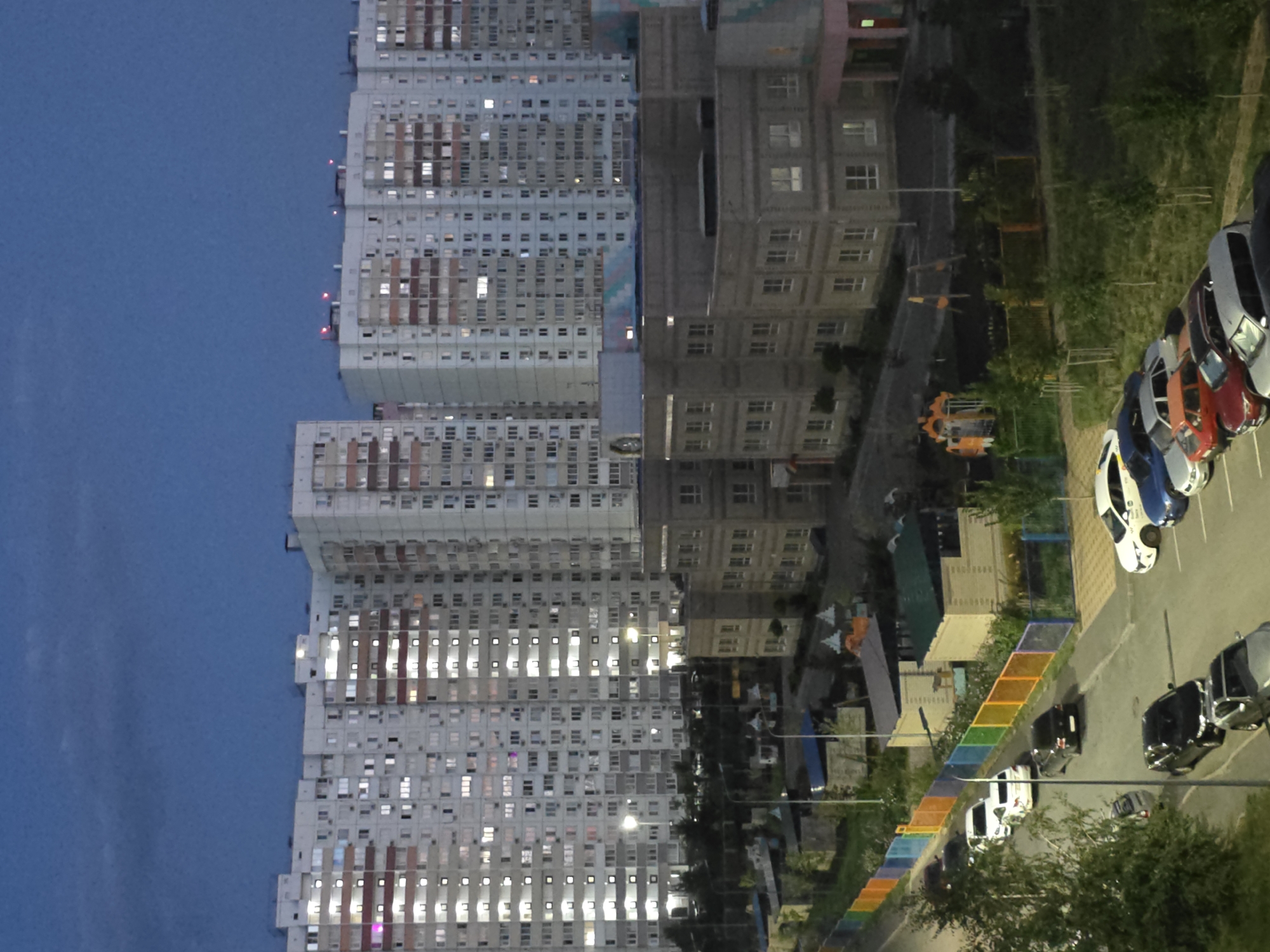 The image depicts a residential complex in Rostov-on-Don, Russia, at dusk. In the background, several tall, multi-story apartment buildings are illuminated with numerous lit windows. In the middle ground, there are lower-rise school or kindergarten buildings with visible windows and architectural details. In the foreground, a street is lined with parked cars, including white, black, silver, blue, and red vehicles. A colorful fence, composed of alternating orange, yellow, green, blue, and purple panels, runs along the side of the street. Trees and other vegetation are visible throughout the scene. The sky is dark blue, indicating twilight. There are no people clearly visible in the image.