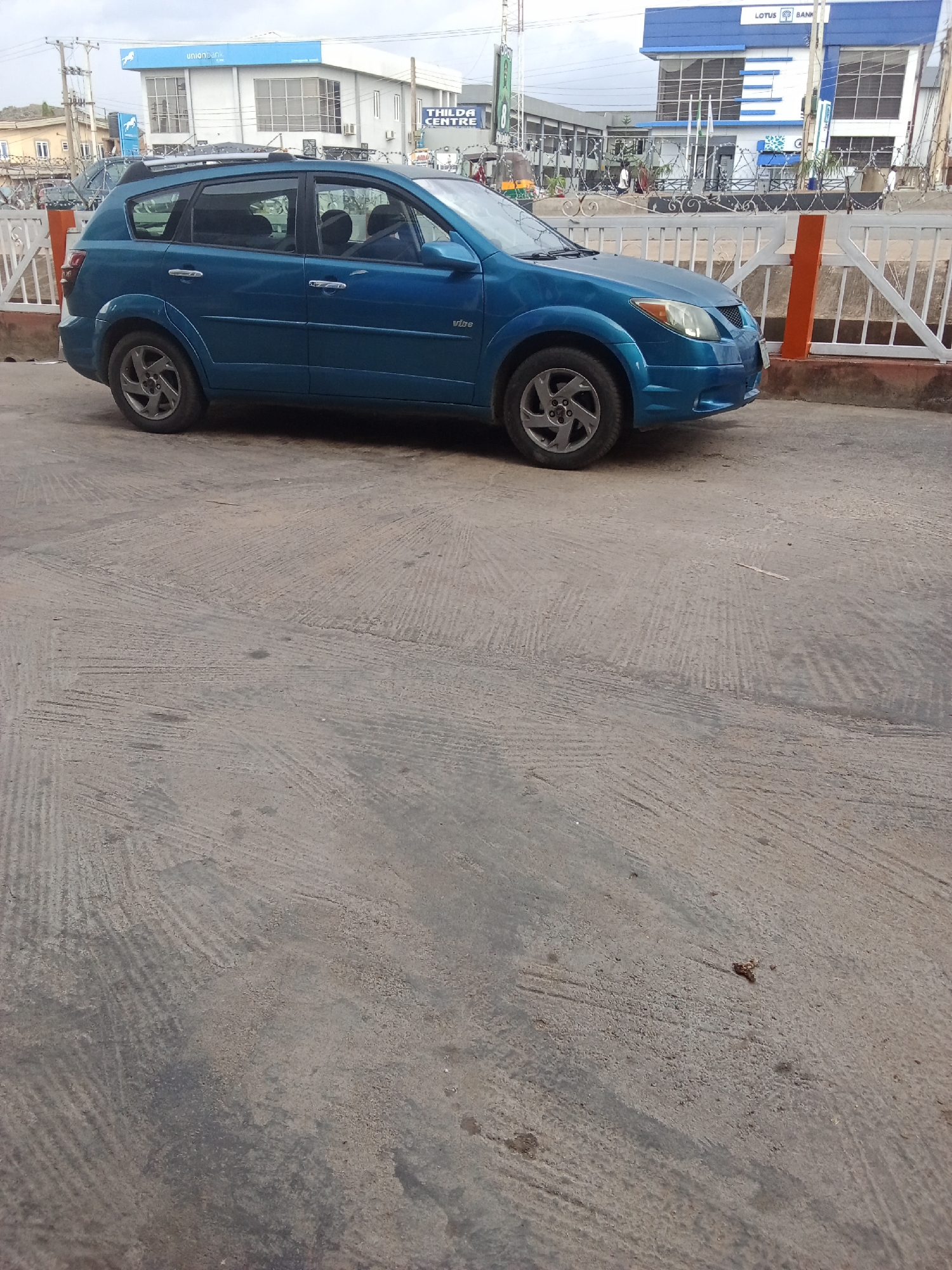 A blue Pontiac Vibe is parked on a concrete surface. The car is facing towards the right of the frame, and its doors are closed. The car's rear is closer to the camera, and the front is further away. Behind the car, a white fence with some barbed wire is visible. Beyond the fence, a building with the text "THILDA CENTRE" can be seen, and further in the background, another building with "LOTUS BANK" signage is present. The environment appears to be an outdoor urban setting, possibly in Jos, Nigeria, given the context. The concrete ground has a rough, textured finish with visible tire marks. The sky is overcast.