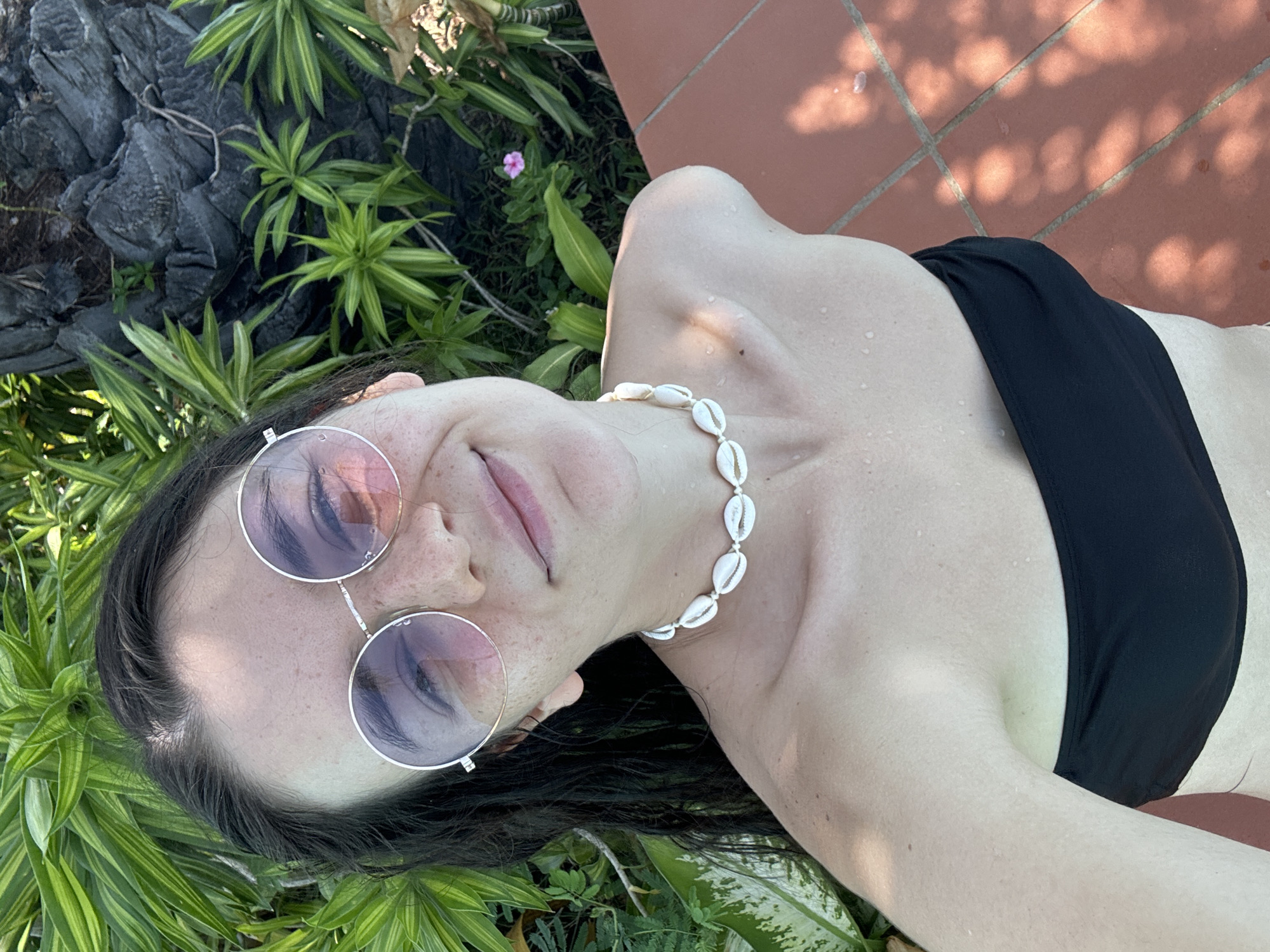 A young woman is seen in a ground-level, angled portrait. She is wearing round, metal-rimmed sunglasses with tinted lenses. She has on a black bikini top, and a necklace composed of white seashells. The woman has droplets of water on her skin. She is surrounded by green foliage on one side, including plants with long, striped leaves. On the other side of her is a red-tiled surface. The city of Phu Thuy Ward, Vietnam, is unspecified in the image.