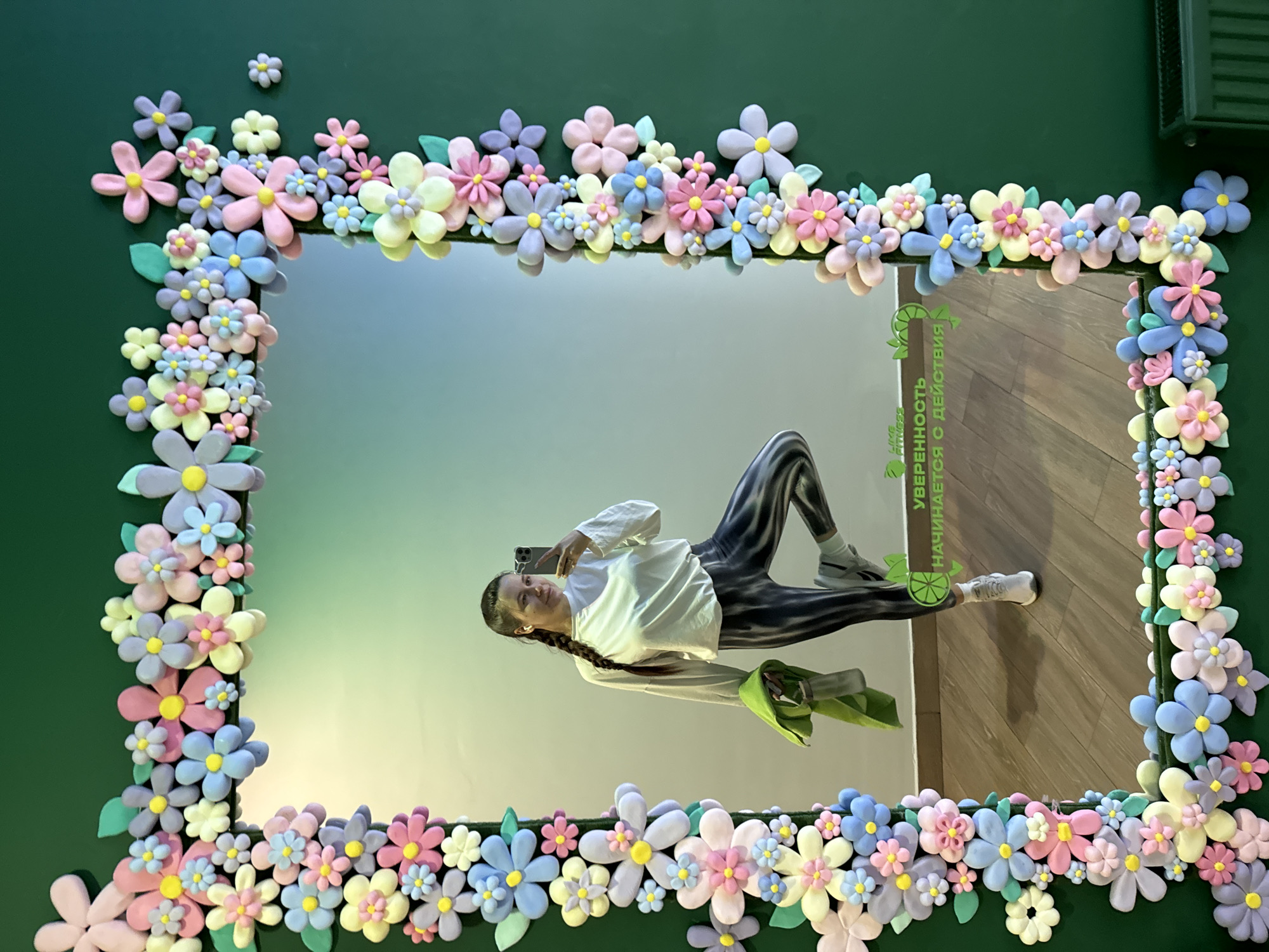A vertical shot captures a woman taking a mirror selfie, framed by a vibrant, decorative border. The mirror itself is set against a dark green wall. The frame is elaborately adorned with numerous pastel-colored, three-dimensional flower-shaped decorations in shades of pink, blue, purple, yellow, and light green, interspersed with small green leaves. These decorations appear to be made from a soft material, possibly clay or foam.
Reflected in the mirror, a young woman is seen holding a dark-colored smartphone to take a selfie. She is wearing a white long-sleeved top, dark patterned leggings, and white athletic shoes, clearly displaying the Reebok logo on one. Her dark hair is styled in a braid. In her right hand, she holds a bright green fabric item, possibly a towel or clothing, and a transparent bottle or cup. The woman's reflection is rotated approximately 90 degrees clockwise, making her appear to be lying down on the reflected background. Her expression is neutral or displays a slight smile.
The reflected background behind the woman shows a light-colored wall and a wooden floor, indicating an indoor setting, likely a fitness center. This is further supported by visible text vertically oriented on the right side of the mirror, which reads in Russian: "УВЕРЕННОСТЬ НАЧИНАЕТСЯ С ДЕЙСТВИЯ" (Confidence begins with action). Below this, part of a logo "LIKE FITNESS" is visible, along with a circular green emblem resembling a lime or lemon. The bright and even lighting suggests an artificial indoor light source. The photo is taken in Vniissok, Russia.