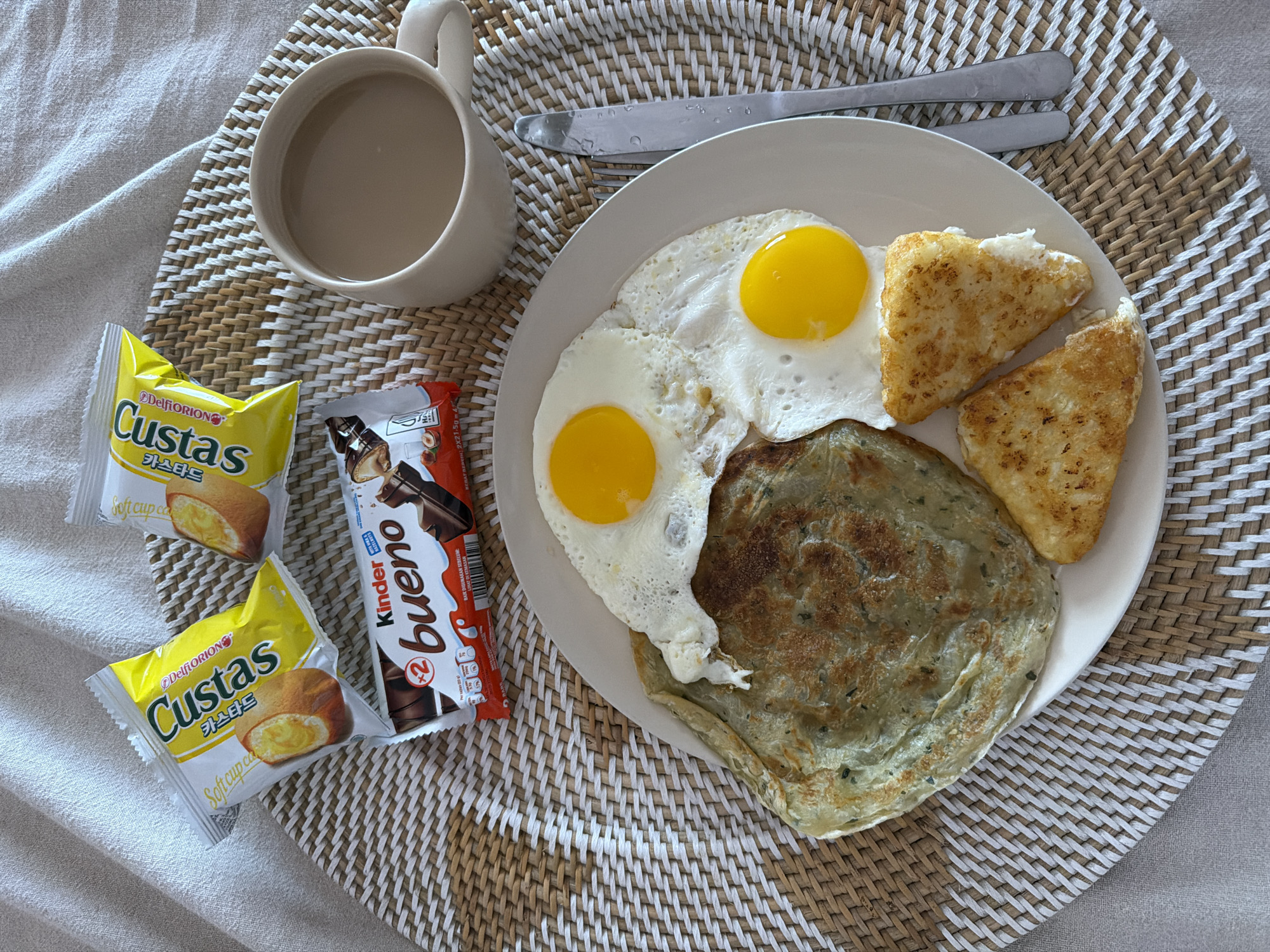 The image is a top-down shot of a breakfast scene. A white plate holds two sunny-side-up eggs, two triangular hash browns, and a round flatbread with visible green specks, possibly herbs. A silver knife rests above the plate on a woven placemat. To the left, there's a mug filled with a beige liquid, likely coffee or tea, along with two yellow packages labeled "Custas" and a "Kinder Bueno" chocolate bar. The background features a soft, neutral-colored fabric, suggesting a casual setting. Based on the "BAIK DIGUNAKAN SEBELUM" text on the Kinder Bueno packaging, the image may have originated in North Kuta, Indonesia.