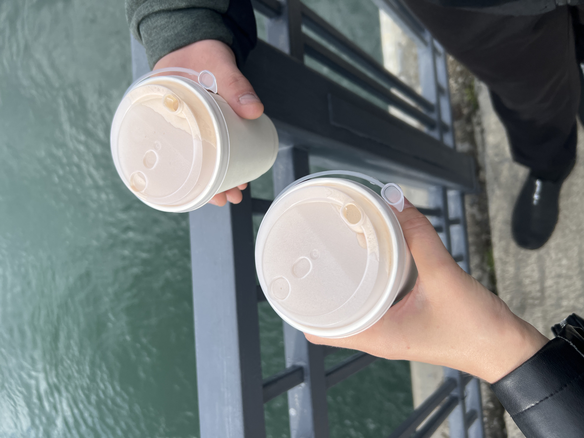 Two individuals are holding coffee cups with plastic lids atop a bridge overlooking a body of water. One person wears a green sweater while the other wears a black jacket. The bridge has a dark gray metal railing. A black dress shoe is partially visible in the upper right corner of the photo. The location may be Irkutsk, Russia.