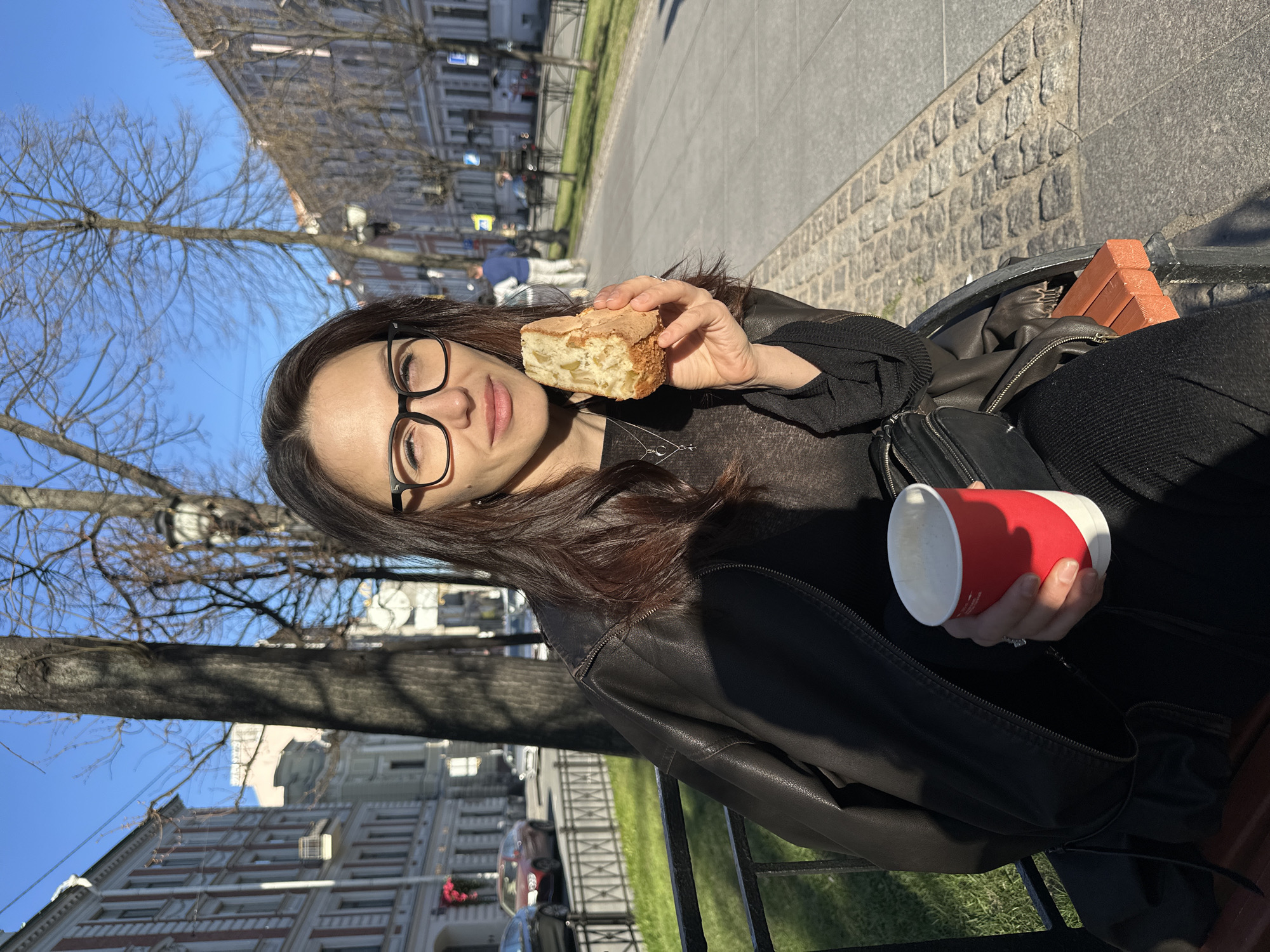 A woman is sitting on a bench in Saint Petersburg, Russia, holding a piece of cake in her left hand and a red and white paper cup in her right. She is wearing glasses, a black jacket, and dark clothing. A black purse rests on the bench next to her. The bench is located on a sidewalk next to a grassy area and a street. Buildings line the opposite side of the street, and bare trees are visible in the background. The weather appears sunny. The cobblestone pavement is visible.