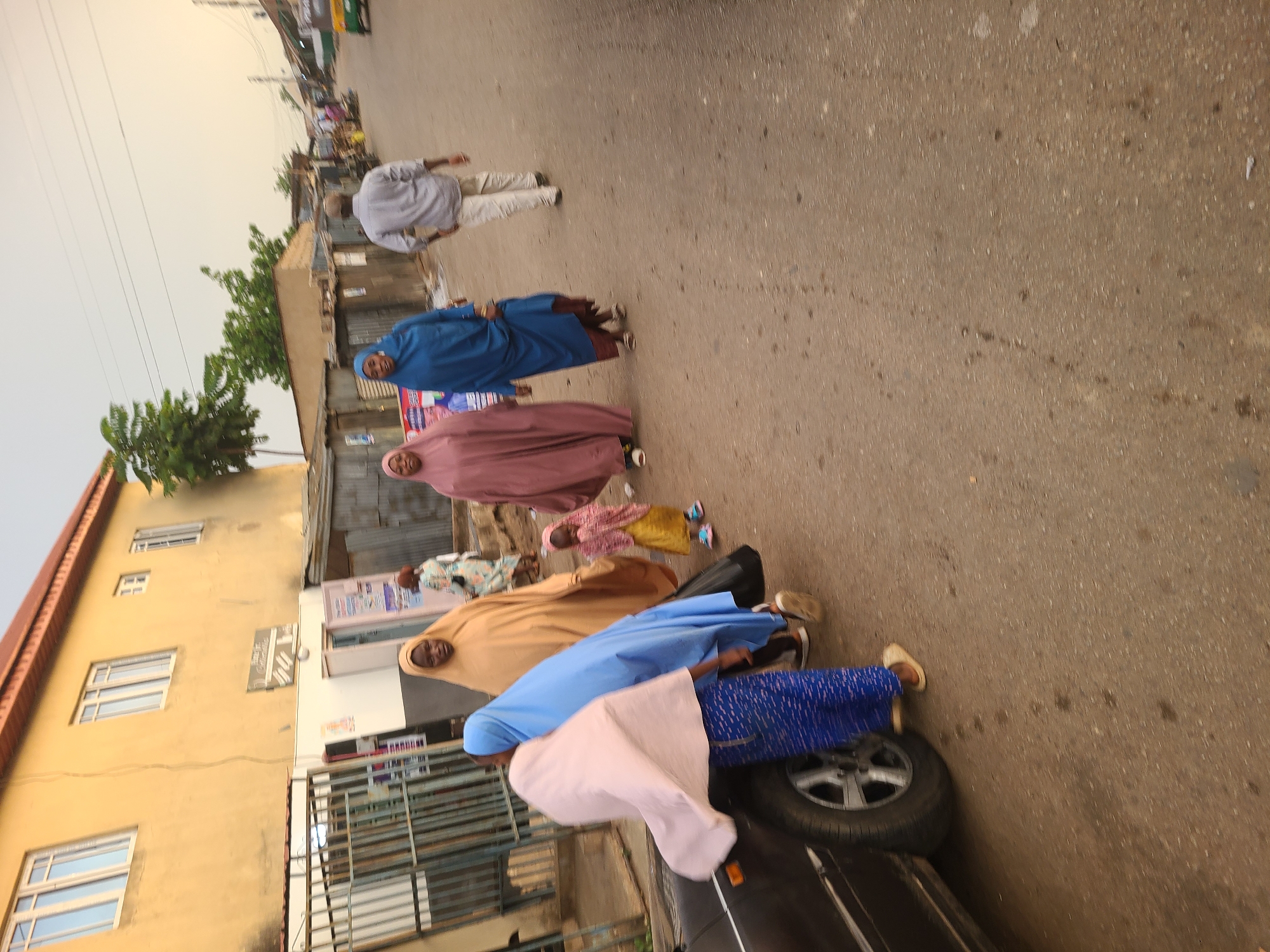 The image shows a street scene in Jos, Nigeria. Several people are walking along the road. Most of the women are wearing traditional Muslim clothing, including headscarves and long robes. There is a building on the left side of the frame. A black car is parked on the right side of the frame. The road is paved. There are trees and power lines visible in the distance.