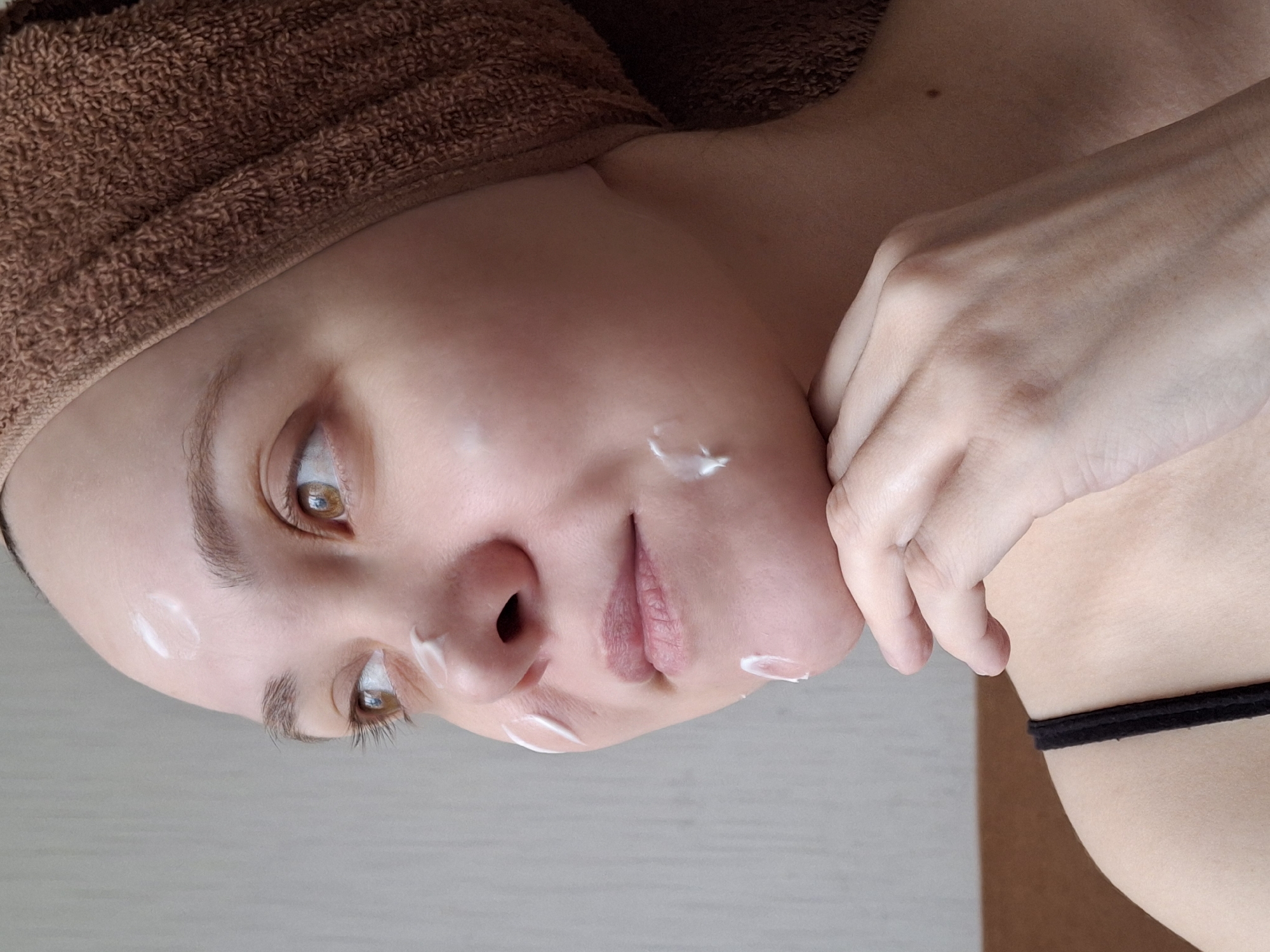 A person is applying face cream. The person has a brown towel wrapped around their head. There are small dabs of white cream on their forehead, cheek, nose, and chin. Their skin is fair. The person has brown eyes and light brown eyebrows. Their right hand is near their chin. A black strap is visible on their shoulder. The background is a light grey surface. The image was taken indoors. The location is not discernible from the image. The city of Chaikovsky, Russia, cannot be confirmed as the location.