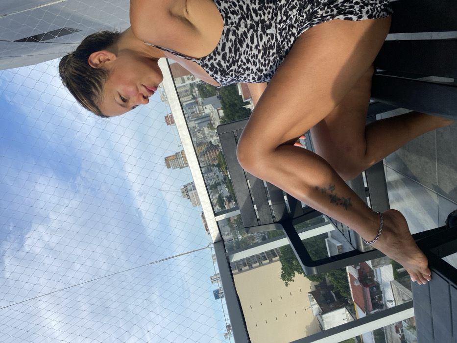 A woman in a black and white patterned romper sits on a chair on a balcony. Her legs are crossed, revealing a tattoo on her lower leg and a silver anklet on her ankle. The backdrop features a cityscape under a blue sky partially covered by a safety net. The woman faces the camera at an angle, showcasing her face and upper body.