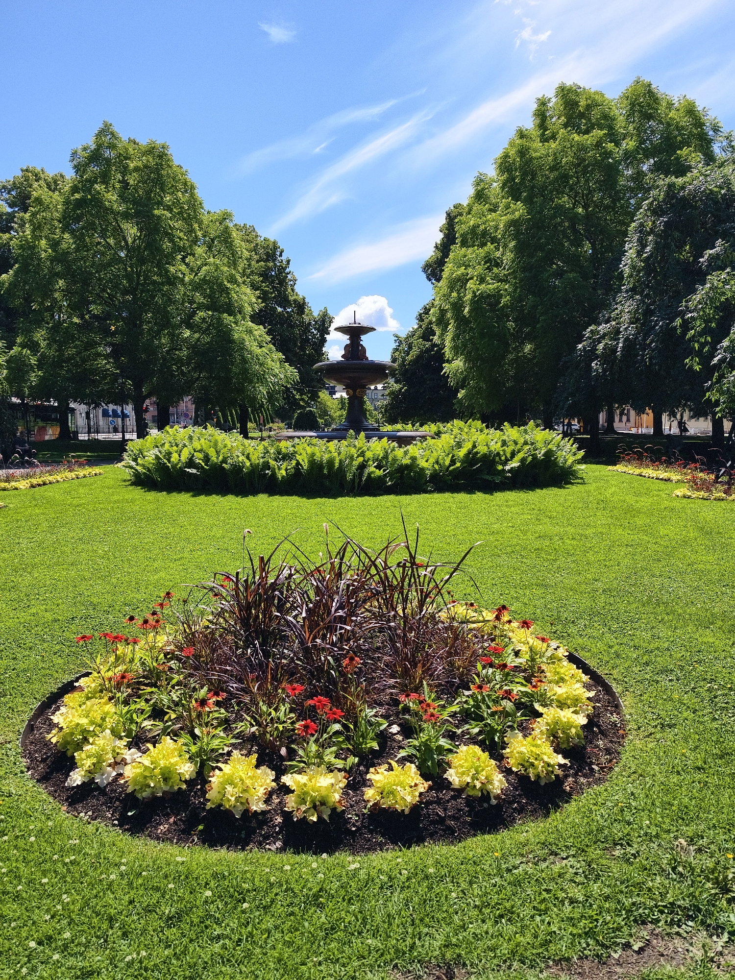 A circular flower bed filled with yellow foliage, red flowers, and dark ornamental grass is in the foreground of a manicured park. Behind it, a dense bed of ferns surrounds a tiered fountain in the middle ground. Rows of trees with vibrant green leaves line the perimeter of the park, and a blue sky with scattered clouds is visible above. The park appears to be located in the city of Norrköping, Sweden.