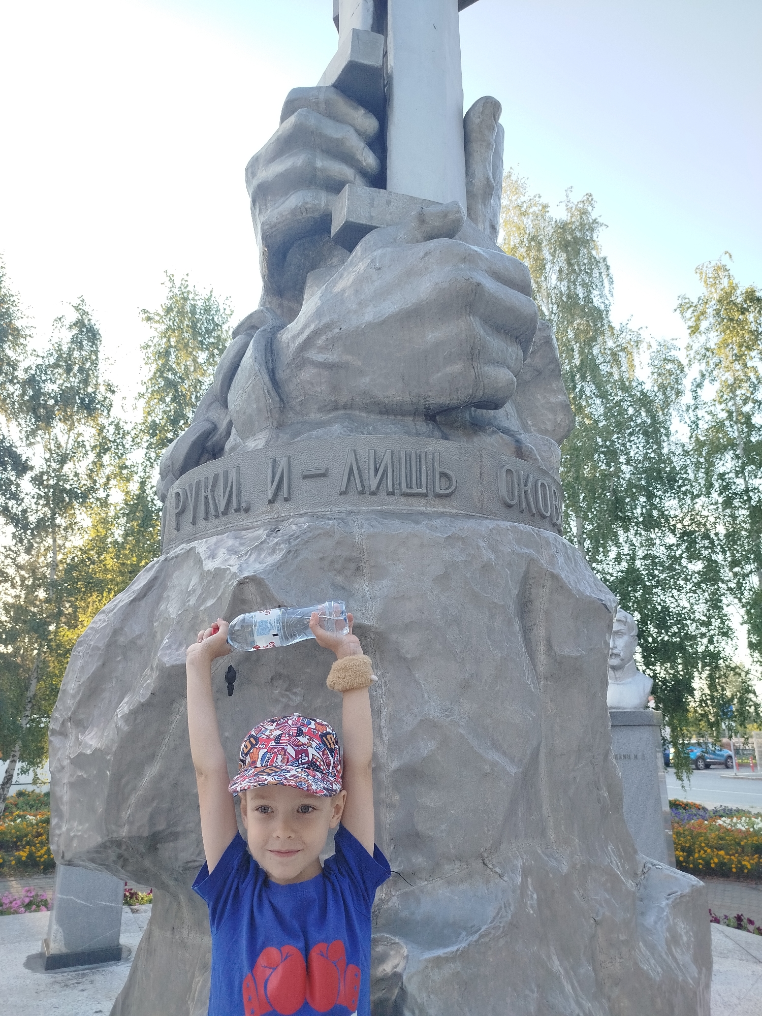 A young boy stands in front of a large stone monument in Yalutorovsk, Russia. The boy is wearing a blue t-shirt with red boxing gloves on it and a colorful baseball cap. He holds a bottle of water above his head with both hands. The monument appears to be made of rough-hewn stone and features large, abstract carved hands. Russian text is visible on a band around the monument. In the background, trees and a bright sky are visible, along with a bust of a man on a pedestal and a blue car driving by. The setting is outdoors, suggesting a public space like a park or square.