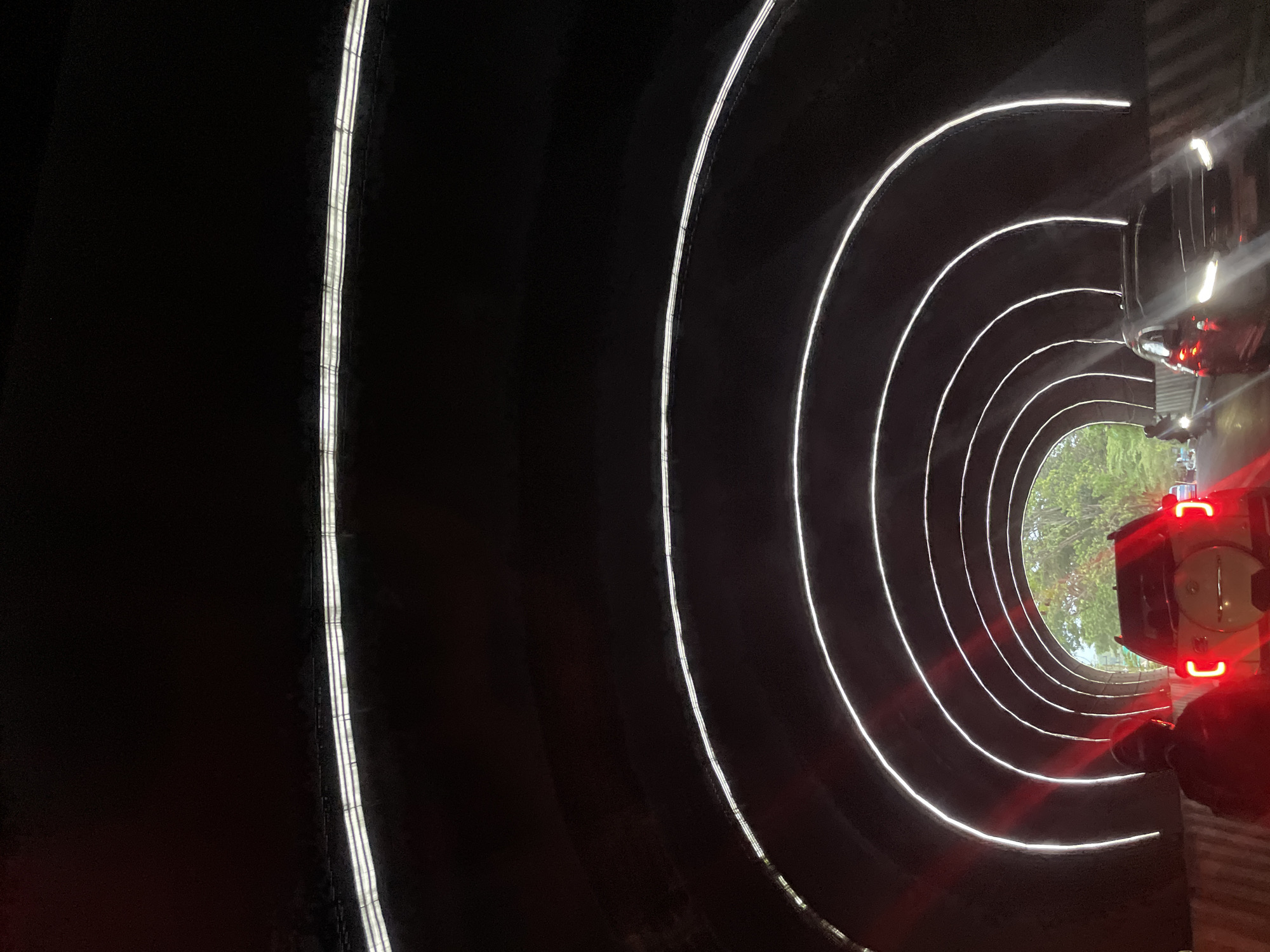 The image depicts the interior of a tunnel in Manado, Indonesia, characterized by a series of illuminated arched structures lining the tunnel walls. Two vehicles are visible: a white SUV in the foreground, facing away from the camera with its brake lights illuminated, and a dark-colored sedan in the right lane, also moving away from the camera. A person on a motorcycle, seen from behind, is in the left lane, partially illuminated by red light. The tunnel's exit is visible in the distance, revealing trees and daylight. The overall scene suggests a moment of traffic within the tunnel.