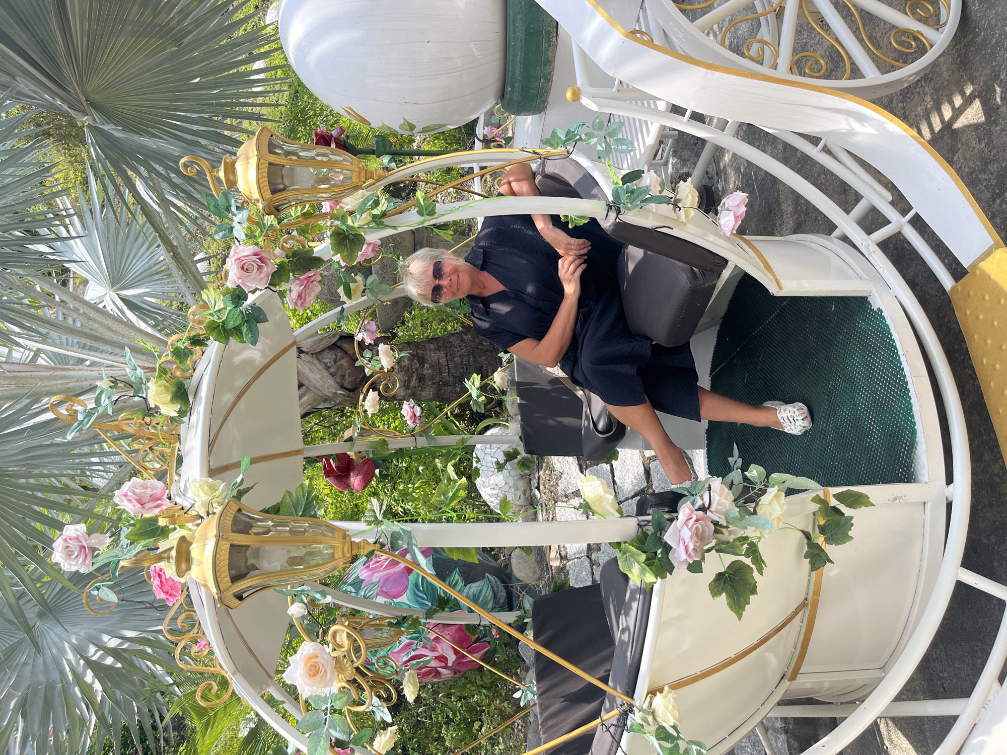 A woman wearing a dark dress and sunglasses sits in a white and gold carriage decorated with flowers. The setting appears to be outdoors in a garden or park, with lush greenery and palm trees visible in the background. The photo may have been taken in Nam Nha Trang Ward, Vietnam, though there is no explicit confirmation from the image itself.