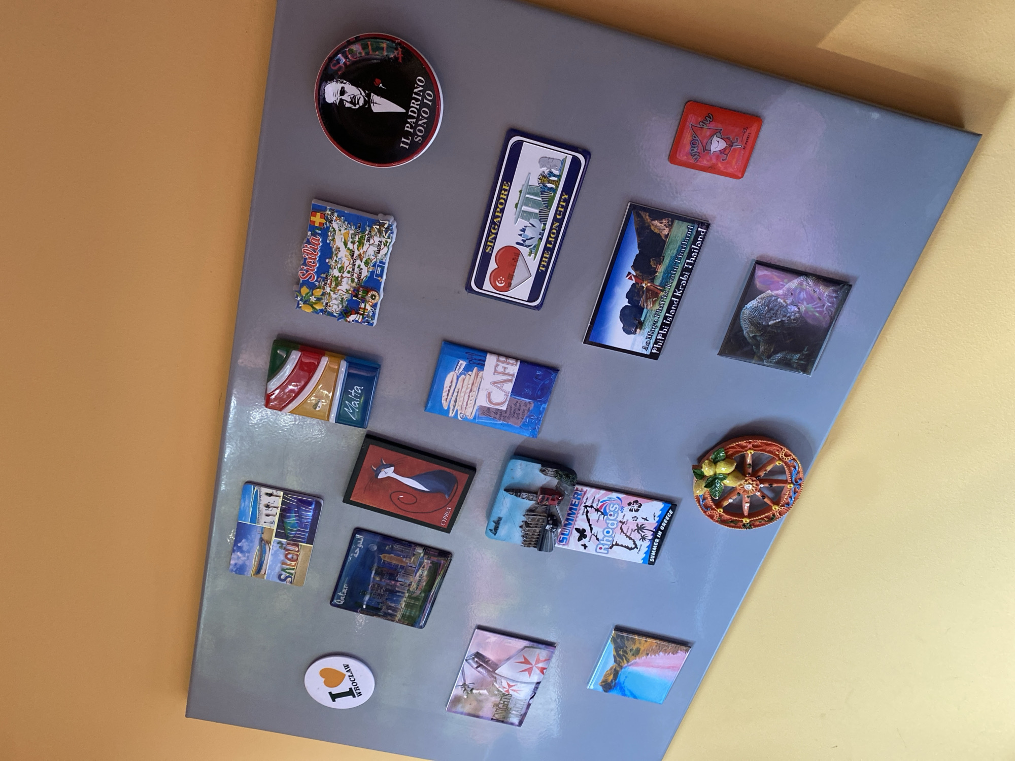 The image shows a collection of refrigerator magnets adhered to a gray magnetic surface, which is mounted on a wall painted a light orange. The magnets represent various travel destinations. Starting from the top left, there is a circular white magnet with "I ♥ WROCŁAW" printed on it in black, with a red heart in place of the word "love." To the right of the "Wrocław" magnet is a square magnet with multiple small images and the text "SALOU" and "SPAIN." Further to the right, a rectangular magnet depicts a scene of a cityscape with the text "Qatar" and Arabic script. Next to the Qatar magnet is a rectangular magnet featuring a black cat with red accents and the text "CYPRUS." Above the Cyprus magnet is a rectangular magnet with the Italian flag colors and the word "MALTA" prominently displayed. To the right of the Malta magnet is a square magnet with a map-like depiction of "Sicilia" (Sicily) with various place names. To the right of the Sicily magnet is a round, dark magnet with a portrait of a man in a suit and the Italian text "IL PADRINO SONO IO." Below the Qatar magnet, on the left, is a square magnet with a white cross on a red background, with the text "KNIGHTS OF MALTA." Below the Knights of Malta magnet is a rectangular magnet showing a coastal landscape with blue water and sandy shores. Below the Cyprus magnet is a rectangular magnet with the word "CAFE" in large letters, superimposed on an image of coffee cups. To the right of the cafe magnet is a rectangular magnet depicting a cityscape with Big Ben and a red double-decker bus, labeled "LONDON" and "SUMMER in GREECE" with "Rhodes" printed in a stylized font. Below the London/Rhodes magnet is a horizontal rectangular magnet showing the iconic skyline of Singapore with the text "SINGAPORE" and "THE LION CITY." Below the Singapore magnet is a horizontal rectangular magnet displaying a tropical island scene with boats, labeled "Ao Nang, Phi Phi Krabi, Thailand. Phi Phi Island, Krabi, Thailand." In the bottom right quadrant, there is a small, red square magnet with a cartoon character and the word "WROCŁAW." To the left of the red Wrocław magnet is a square magnet with a detailed image of a Komodo dragon. At the bottom center, there is a circular, orange magnet with a decorative wheel design and