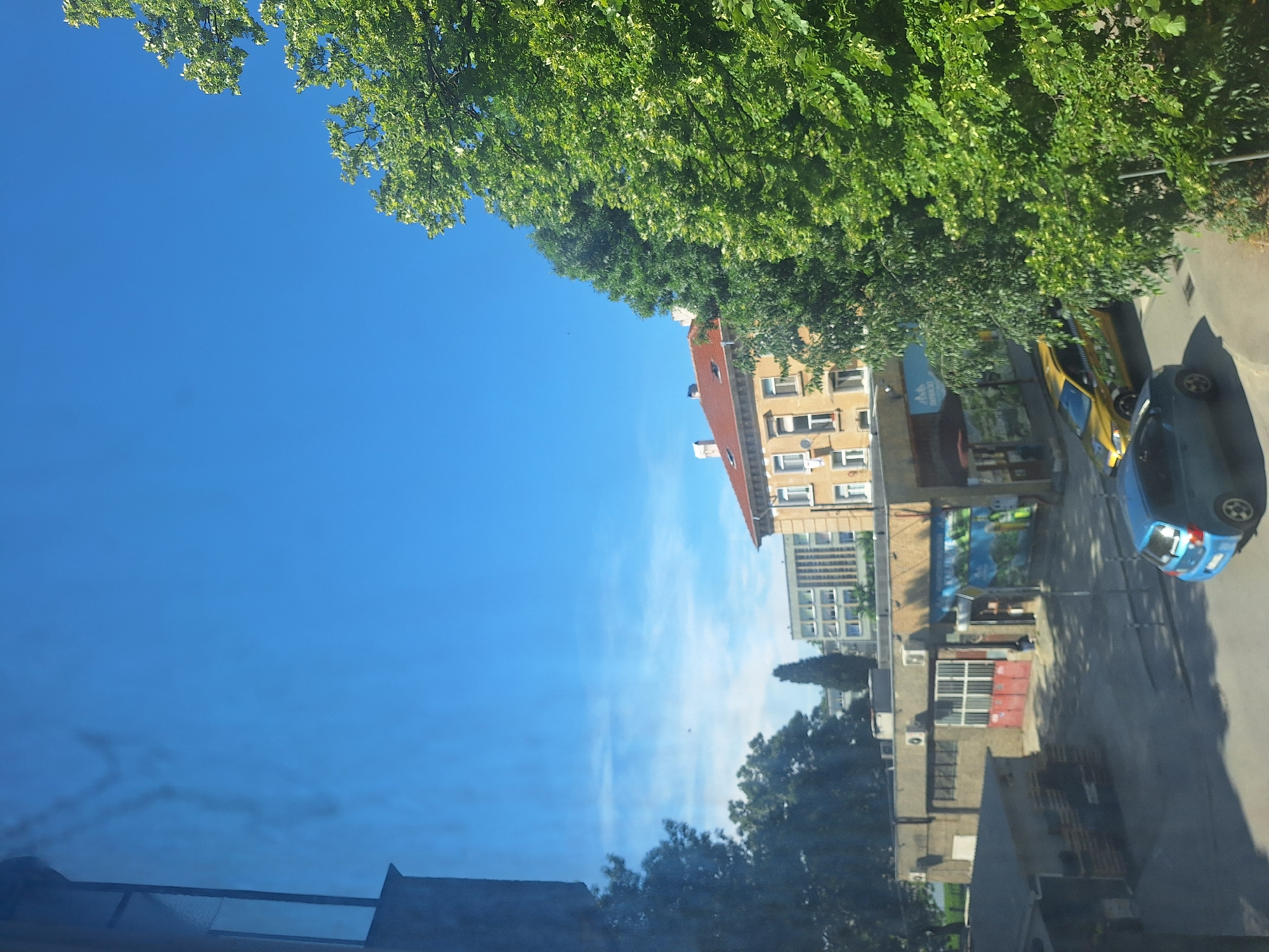 The image depicts an outdoor scene on a bright, clear day in Varna, Bulgaria. In the foreground, a blue car is parked on a paved surface. To the left, a grey building with a dark roof is partially visible. Behind the blue car, a yellow taxi is parked on the asphalt. In the mid-ground, a low-rise commercial building with a light-colored facade and large red doors is prominent. A mural or advertisement is displayed on the side of this building. Further in the background, a taller, light-colored building with multiple windows and a flat roof stands. To the right, lush green trees with dense foliage dominate the scene, their branches reaching towards the clear blue sky. The overall impression is a typical urban environment with elements of infrastructure and nature.