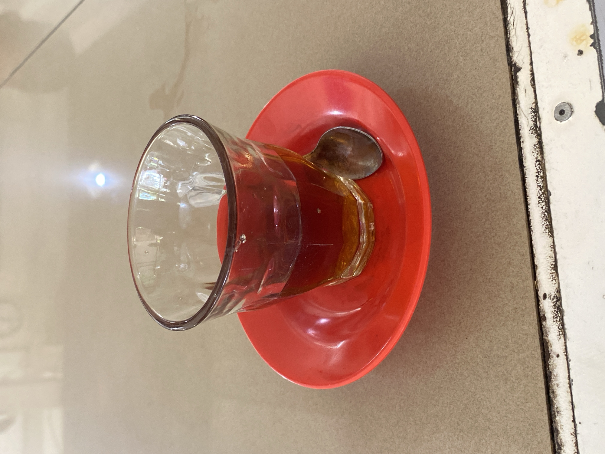 A close-up, eye-level shot shows a glass of tea with a metal spoon on a red saucer. The glass is approximately half full of amber-colored liquid. The saucer is round and red, with a raised rim. The spoon is resting on the saucer to the right of the glass. The scene appears to be set on a light brown, textured surface, possibly a table. In the background, a bright light source is reflected on the surface, and a hint of white furniture is visible. The bottom edge of the frame reveals a white surface with some dark staining and a visible screw. This image was taken in Lhokseumawe, Indonesia.