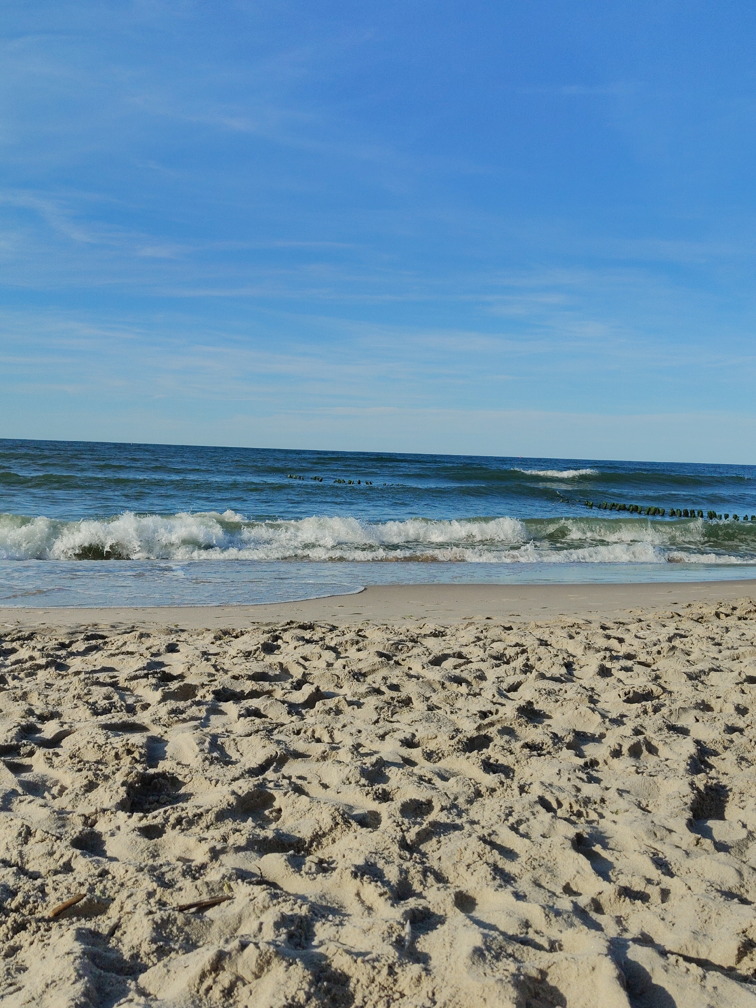 The image shows a sandy beach in Pionersky, Russia, with the Baltic Sea in the background. The sky is blue with some thin, wispy clouds. The sea is a deep blue-green with white-capped waves rolling towards the shore. Along the horizon, there are several wooden groynes extending into the sea. The sand in the foreground is light tan and textured, with some small footprints and debris. There are no people visible in the image. The scene is peaceful and serene, suggesting a calm day at the beach.