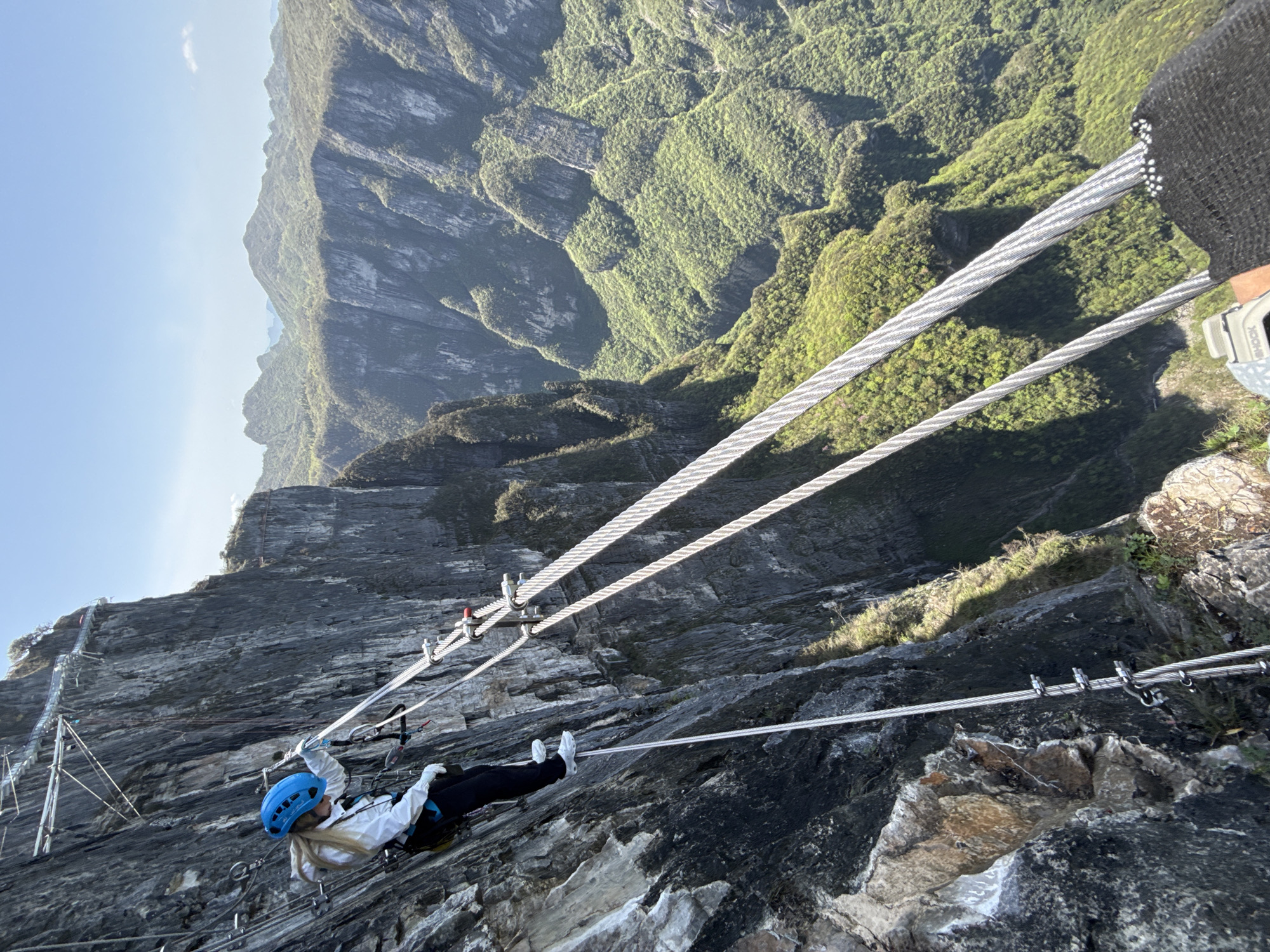 A person wearing a blue helmet, white jacket, black pants, and white shoes is traversing a via ferrata, a protected climbing route, along a steep rock face. The climber is secured to steel cables with a harness and lanyards. The route follows the cliff face, with metal rungs and steps providing support. In the background, there is a mountainous landscape covered in green vegetation. The photograph was taken at Tianmen Mountain in the Yongding District, China.