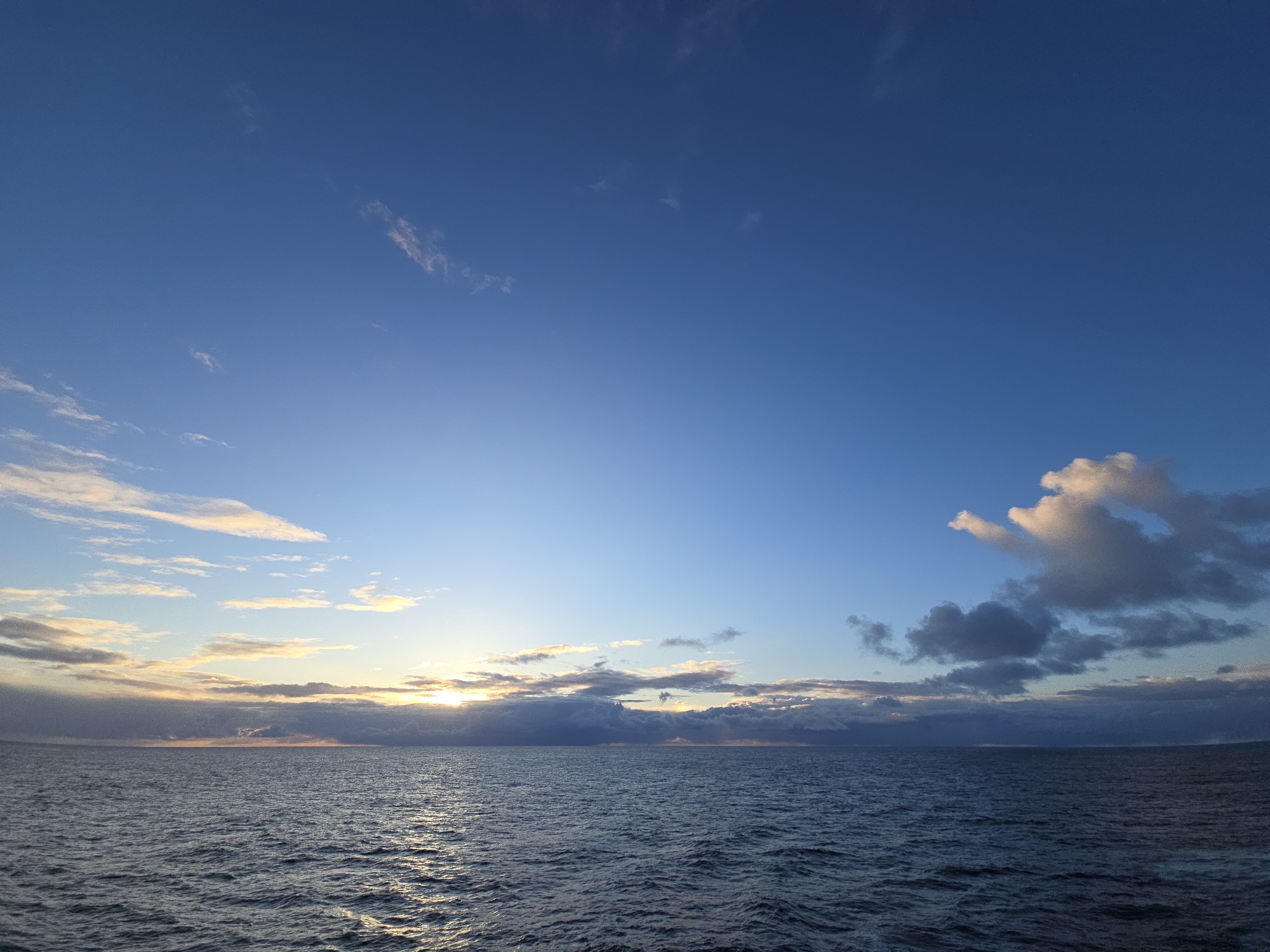 The image depicts a seascape at dawn or dusk. The sky is predominantly blue, transitioning from a deeper hue at the top to a lighter shade near the horizon. Scattered clouds are present, with some tinged with orange and yellow hues from the sun, indicating a sunrise or sunset. The sea is a dark blue, with visible ripples and waves catching the sunlight. The location is Unknown, Unknown.