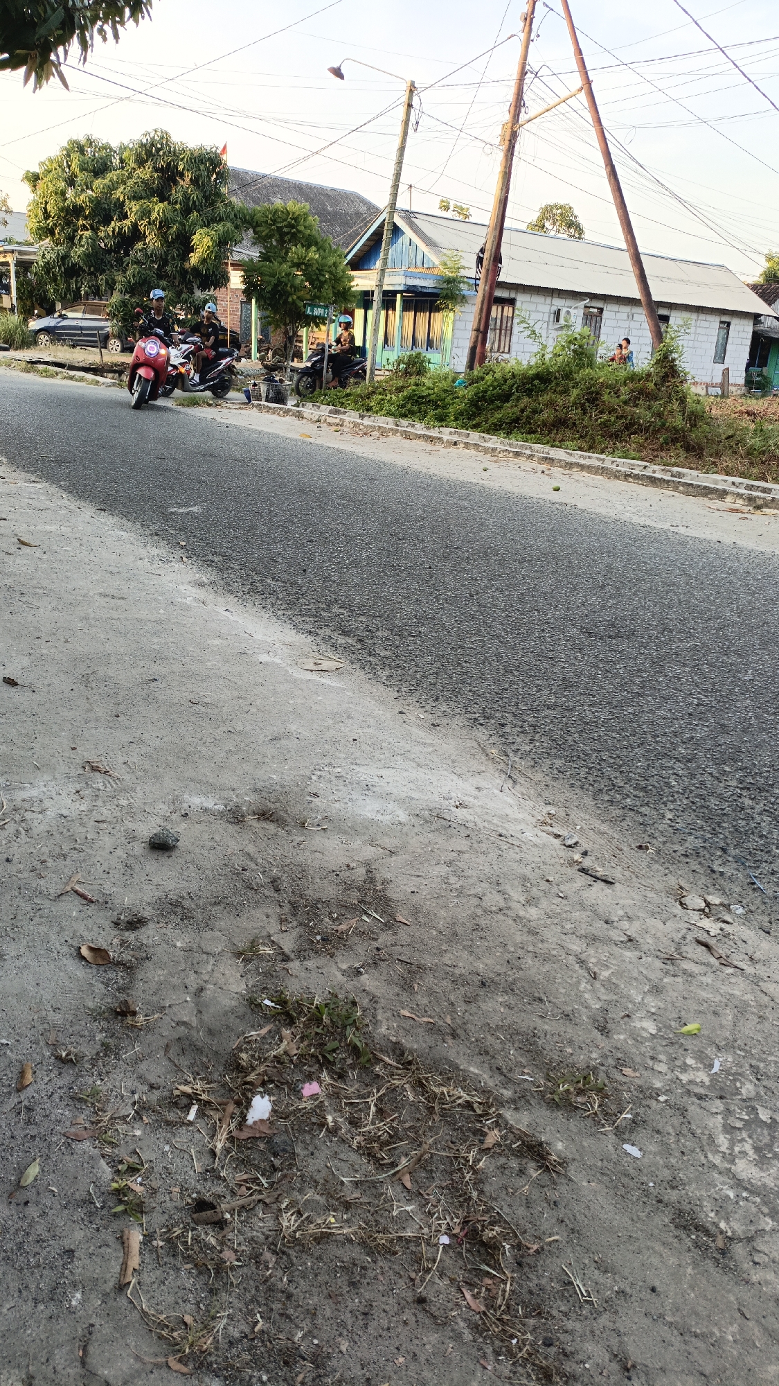 The image depicts a street scene in Gusunge, Indonesia. In the foreground, a grey, rough-textured dirt and concrete roadside is visible, littered with dry grass, leaves, and a few pieces of white and pink litter. The road itself is paved with dark asphalt and runs diagonally across the frame from the bottom left to the upper right.
On the right side of the road, a white, single-story building with a light blue trim is situated behind a dense growth of green bushes. Several utility poles and a network of overhead wires dominate the upper portion of the image, casting shadows.
On the left side of the road, a large tree with lush green foliage stands next to a red motorcycle with a rider wearing a blue helmet and a dark shirt. Behind this, another motorcycle is parked, with two people on it wearing helmets. Further down the road, a dark motorcycle is parked with a person in a uniform and helmet. Two individuals are seen sitting near the building on the right. A dark-colored car is parked on the left side of the road, near the large tree. The overall lighting suggests it is late afternoon or early evening.