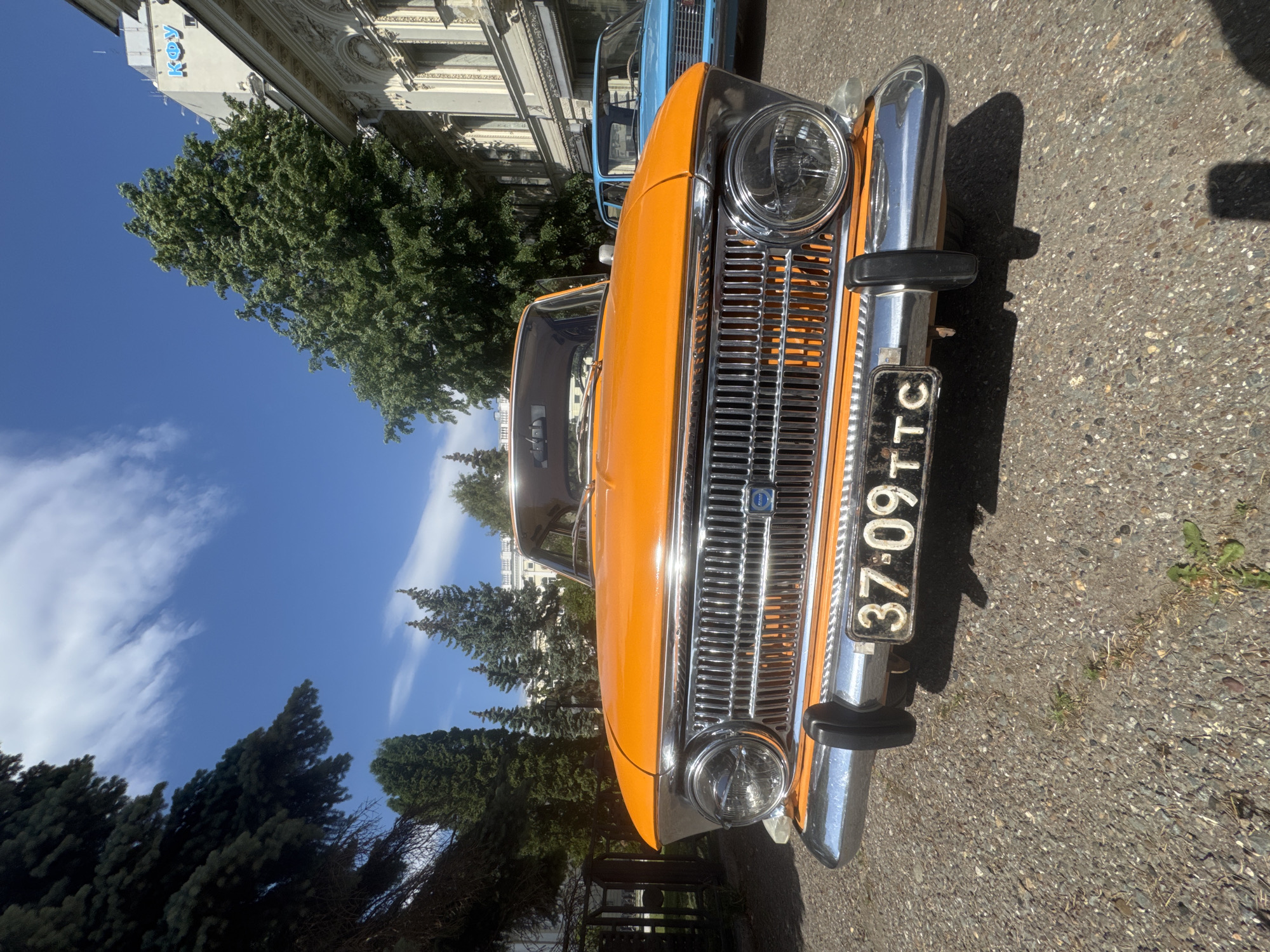 The image shows the front of an orange vintage car with the license plate "37-09 TTC." The car is parked on a street with an asphalt surface. In the background, there are green trees, a building with ornate architectural details, and a partly cloudy blue sky. Based on the architectural style of the building in the background, the location is in Kazan, Russia. Another blue vintage car is partially visible behind the orange car.
