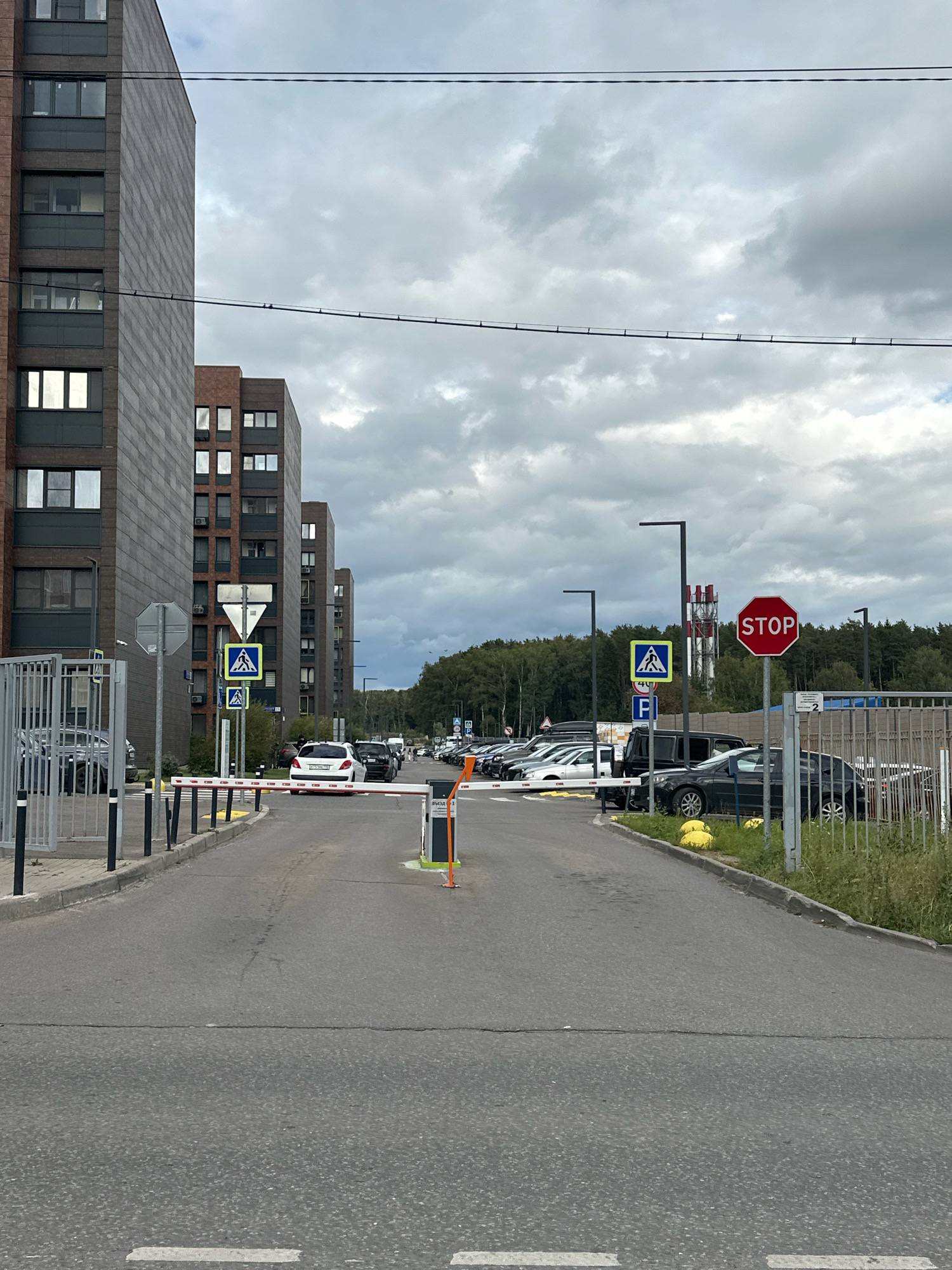 The image depicts an outdoor scene in the city of Vidnoye, Russia, with a view of residential buildings and a parking area. In the foreground, an asphalt road leads towards a white barrier arm that is partially lowered, indicating an entry or exit point for vehicles. To the left of the road, a metal fence with vertical bars borders a sidewalk and a grassy area. Several pedestrian crossing signs with yellow triangular backgrounds and white pedestrian silhouettes are visible along the road.
Dominating the left side of the image are tall, multi-story residential buildings with a brick facade. The buildings feature numerous windows and air conditioning units. On the right side of the road, a row of parked cars is visible, extending into the background. A large red octagonal STOP sign is prominently displayed on a pole, accompanied by a blue rectangular sign with a white letter "P" for parking. In the far distance, a dense line of trees forms the horizon under a cloudy sky. The lighting suggests it is daytime, possibly overcast.