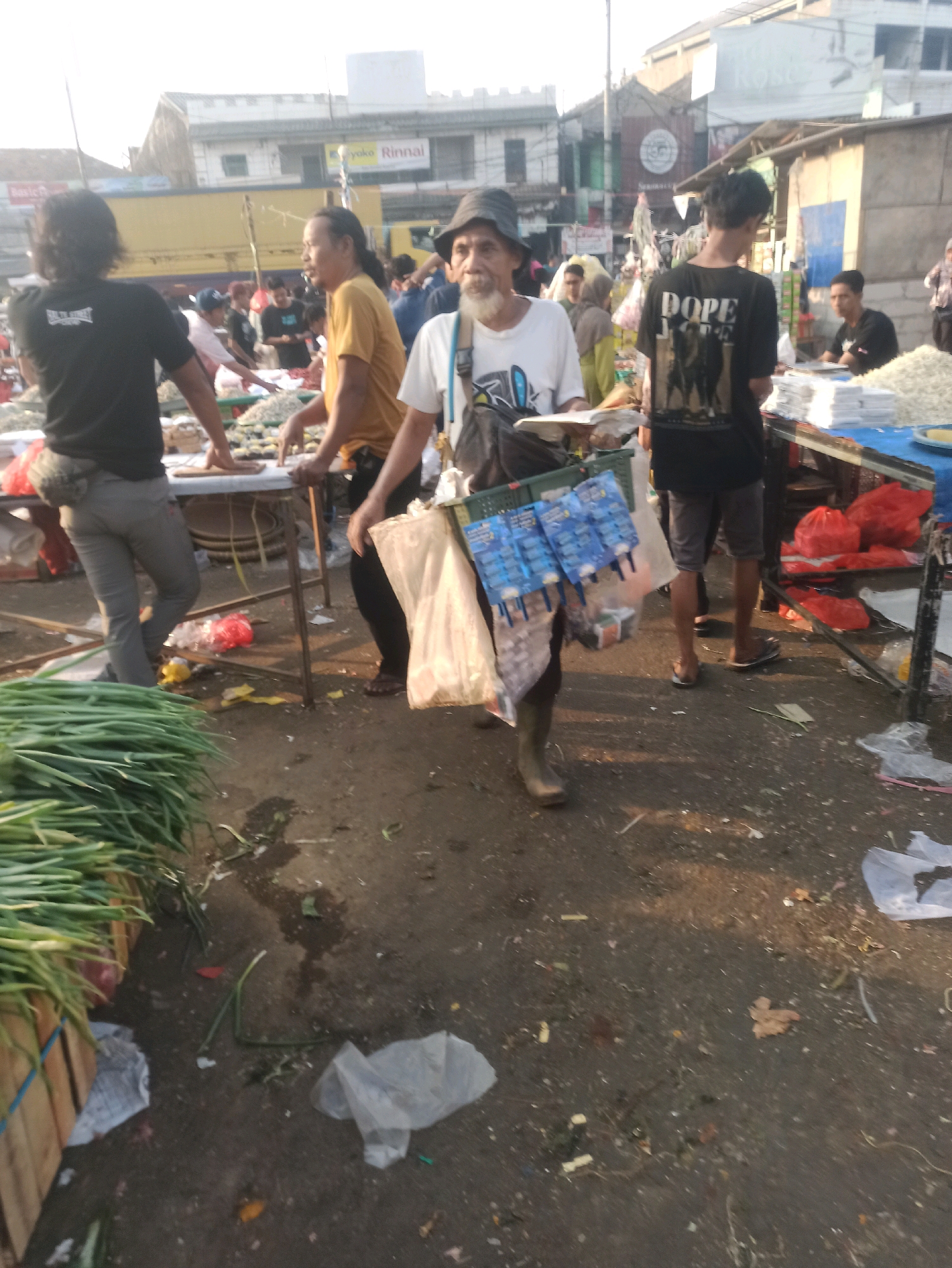 The image depicts a busy outdoor market scene, likely in Cikarang, Indonesia. The ground is paved with dirt and debris, indicative of a well-trodden marketplace. In the foreground, a large bundle of green onions is neatly stacked on the left, suggesting fresh produce is being sold.
The central figure of the image is a man wearing a dark hat and a white t-shirt, carrying a basket filled with various packaged goods, including what appear to be small sachets of candy or snacks, displayed on the front of the basket. He is also holding some food items in his left hand. He wears dark pants and rubber boots. To his left, a person in a black t-shirt and grey pants stands behind a table, with their back mostly to the camera. To the man's right, another individual in a black t-shirt with the word "DOPE" on the back is visible, wearing shorts and sandals.
In the background, several other people are present, some appearing to be vendors or shoppers, browsing at stalls. Buildings, including one with "yoko Rinnai" signage, are visible in the distance, indicating an urban or semi-urban setting. The lighting suggests it is daytime, possibly late afternoon due to the long shadows and warm light. The overall atmosphere is one of commerce and daily life in a local market.
