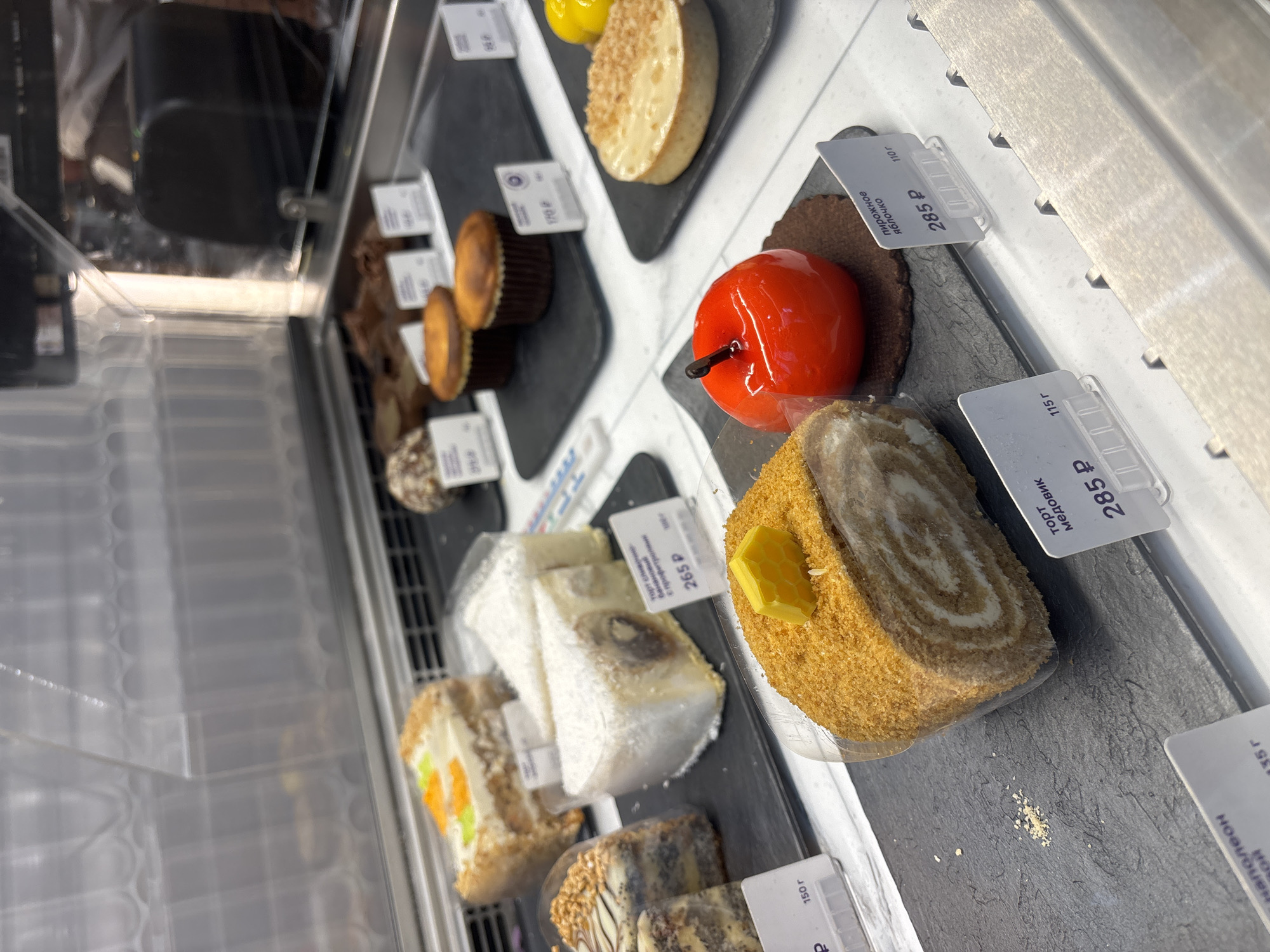 Here is a description of the image:
The image shows a display case filled with various cakes and pastries. A bright red, apple-shaped pastry with a black stem is featured near the center of the frame. Next to it is a roll cake with a yellow honeycomb-shaped piece on top. Slices of other cakes, some with white frosting and fruit pieces, are also visible. Price tags in Russian currency (₽ - Rubles) are displayed next to each item, indicating the cost per weight unit. The price tags read "285 P", "265 P", and "1702" among other unreadable labels. The environment is a well-lit display case, suggesting a bakery, cafe, or similar establishment, possibly located in Saint Petersburg, Russia, based on the Cyrillic alphabet used on the price tags. The items are arranged on slate serving dishes.