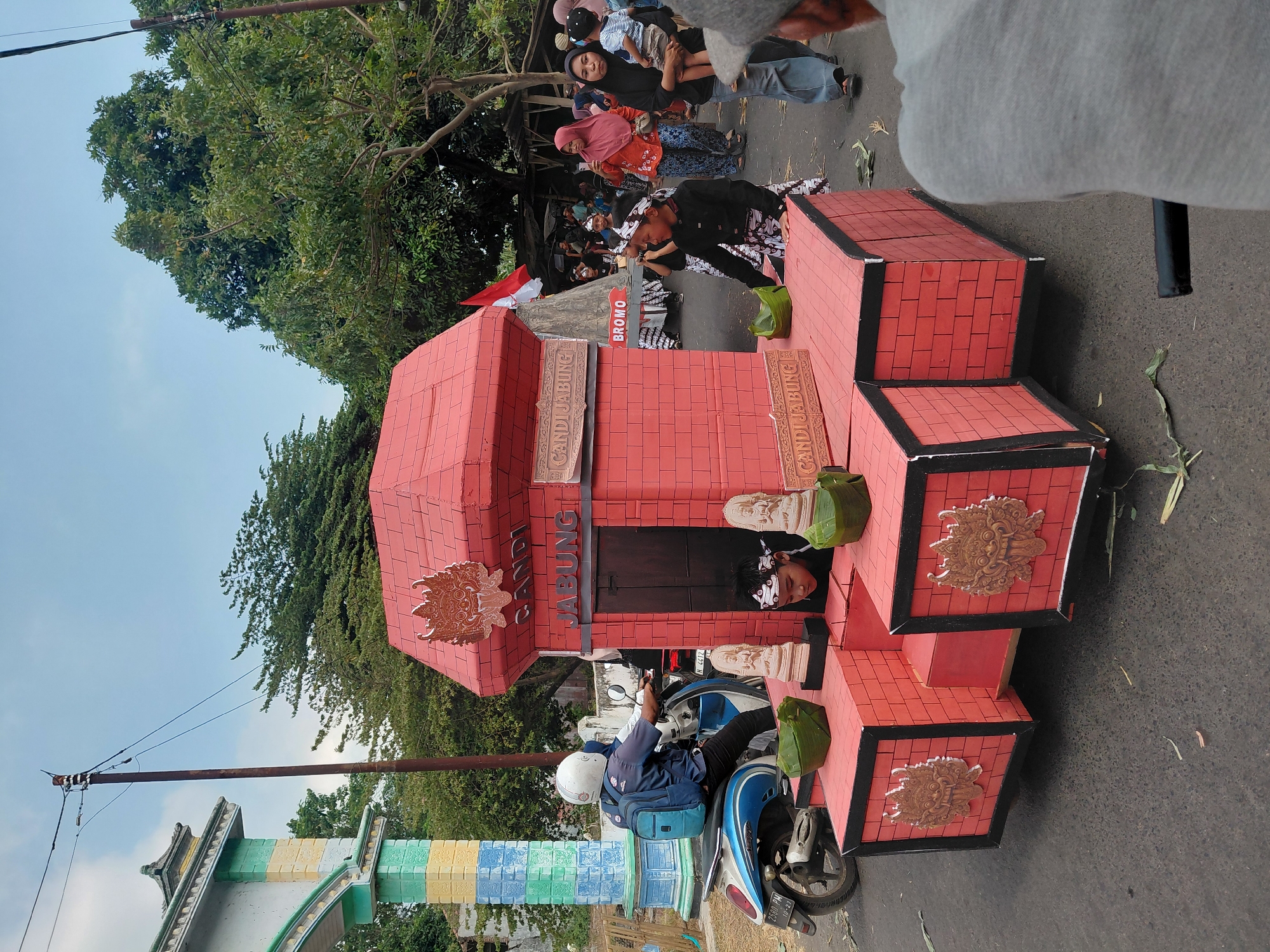 This is a medium shot, taken outdoors during daylight, capturing a parade float in Bimo, Indonesia. The float is designed to resemble a red brick building with a red tiled roof, adorned with decorative carvings. The words "CANDI JABUNG" are displayed on the float in two locations. Two children, one wearing a traditional patterned garment and headband, are positioned on the lower level of the float. Another child is partially visible inside the structure.
To the left of the float, a person wearing a white helmet and a blue backpack is riding a blue and white scooter. In the background, a colorful arched gateway stands to the left, with trees and a utility pole visible behind it. Further back, a crowd of people are observing the parade, some holding cameras. The ground is paved with asphalt, and scattered leaves are present. The overall atmosphere suggests a cultural event or celebration.