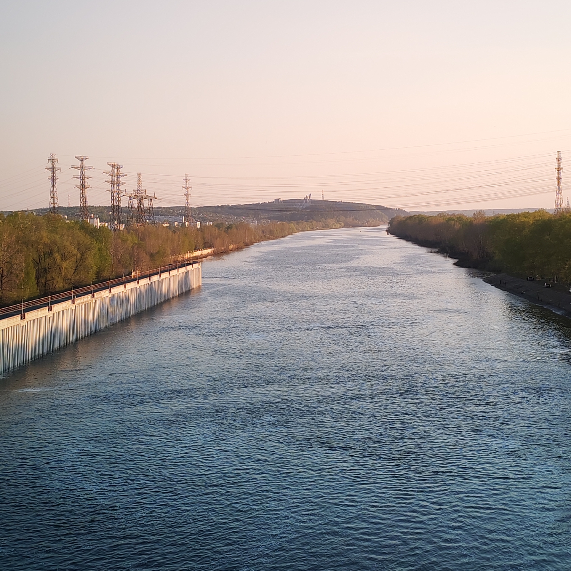 The image shows a wide river flowing through the city of Chaikovsky, Russia. The river appears calm with gentle ripples across its surface. On the left bank, there is a long concrete structure, possibly a pier or a protective barrier, running parallel to the river. Trees line both banks, indicating a green space along the river. In the background, a hill can be seen with structures on top, perhaps a monument or industrial complex. Power lines and electrical towers traverse the scene, suggesting an urban or industrial setting. On the right bank, near the water's edge, there are some indistinct dark shapes that might be people or objects on a beach. The sky is light, indicating either early morning or late evening, with a soft, diffused light.