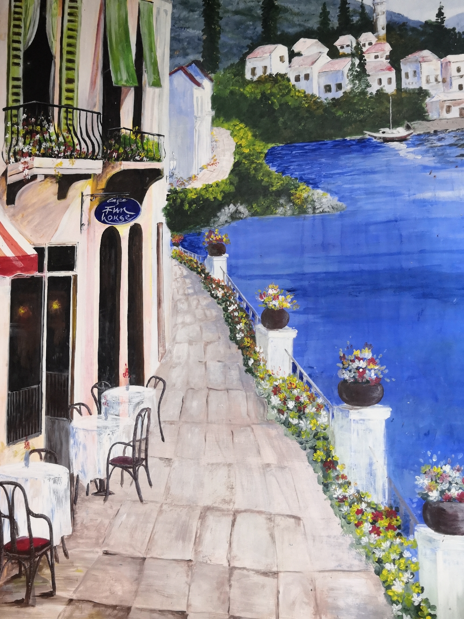This is a painting of a waterfront cafe with tables set outside. The cafe has a sign that says "cafe Fun House". In the background, there are white buildings with red roofs, lush green trees, and a blue body of water with a sailboat. The walkway leading to the cafe is paved with stones and lined with potted flowers. The painting style is impressionistic with bold brushstrokes and vibrant colors. The specific location in Nha Trang Ward, Vietnam is not depicted or identifiable in the painting.