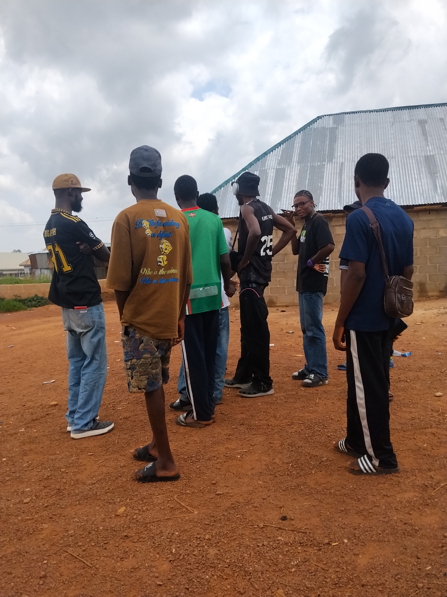 A group of seven young men are gathered outdoors in Durumi, Nigeria, under a cloudy sky. They are standing on a reddish-brown dirt ground covered in small stones and scattered debris. Behind them, a corrugated metal building with a green trim runs across the background, and a brick wall is visible to its right.
The men are dressed in casual attire. One man in the foreground on the left wears a black jersey with yellow lettering and the number '31', paired with light blue jeans. To his right, another man is seen from the back, wearing a tan t-shirt with graphics and camouflage shorts. A third man, also from the back, is in a bright green t-shirt with a small flag emblem. Further to the right, a man in a black t-shirt with text is talking on a phone. Another man in a black sleeveless shirt with 'GAFORO' and '25' printed on it is also present. Two men on the far right are wearing blue and dark t-shirts respectively, with one carrying a brown satchel. Their stances and gazes suggest they are engaged in conversation.